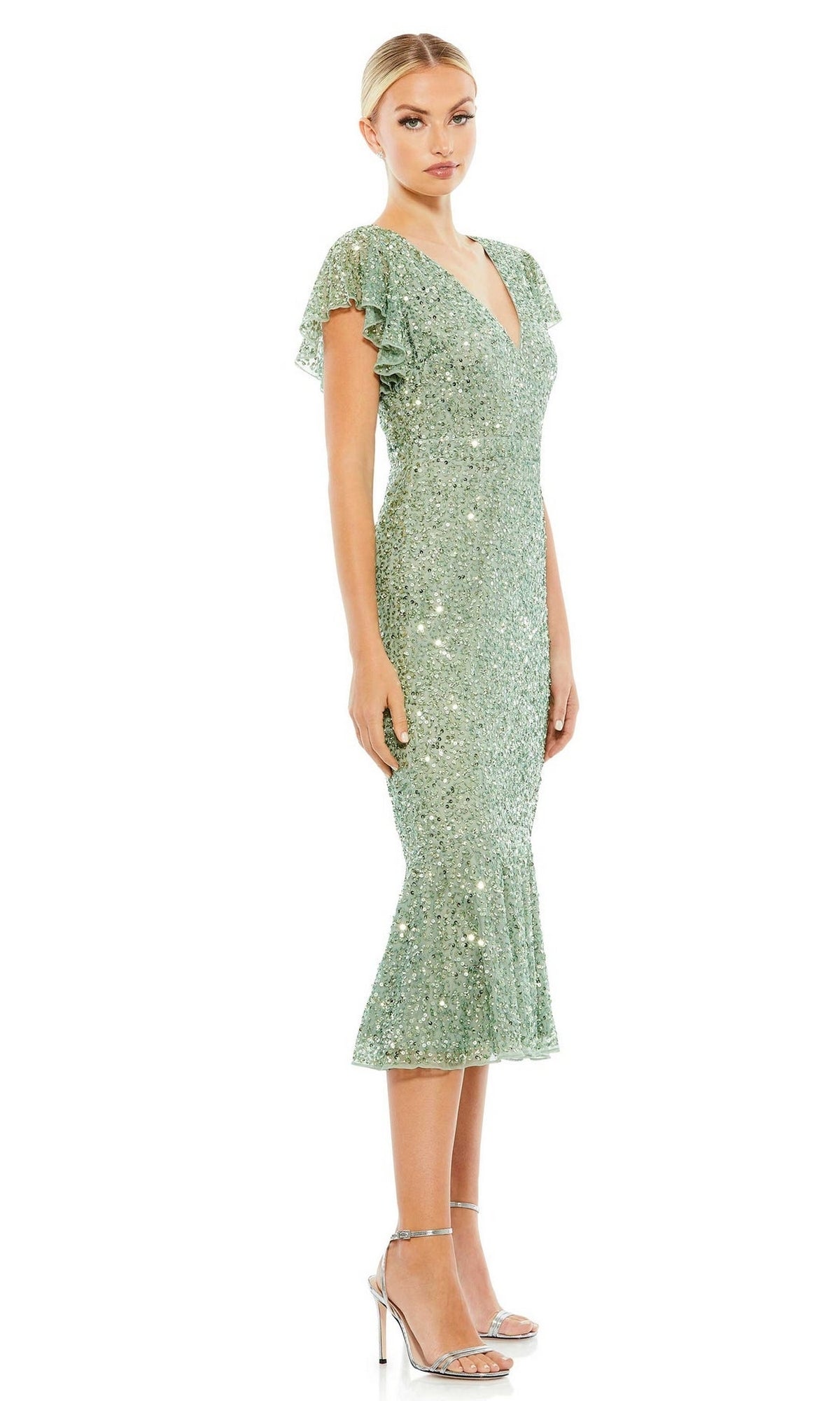 A woman models the Short Wedding Guest Dress 10801 by Mac Duggal, a sparkly sage green sequin trumpet dress with short sleeves, V-neck, and flared hem, paired with silver heels against a white backdrop.