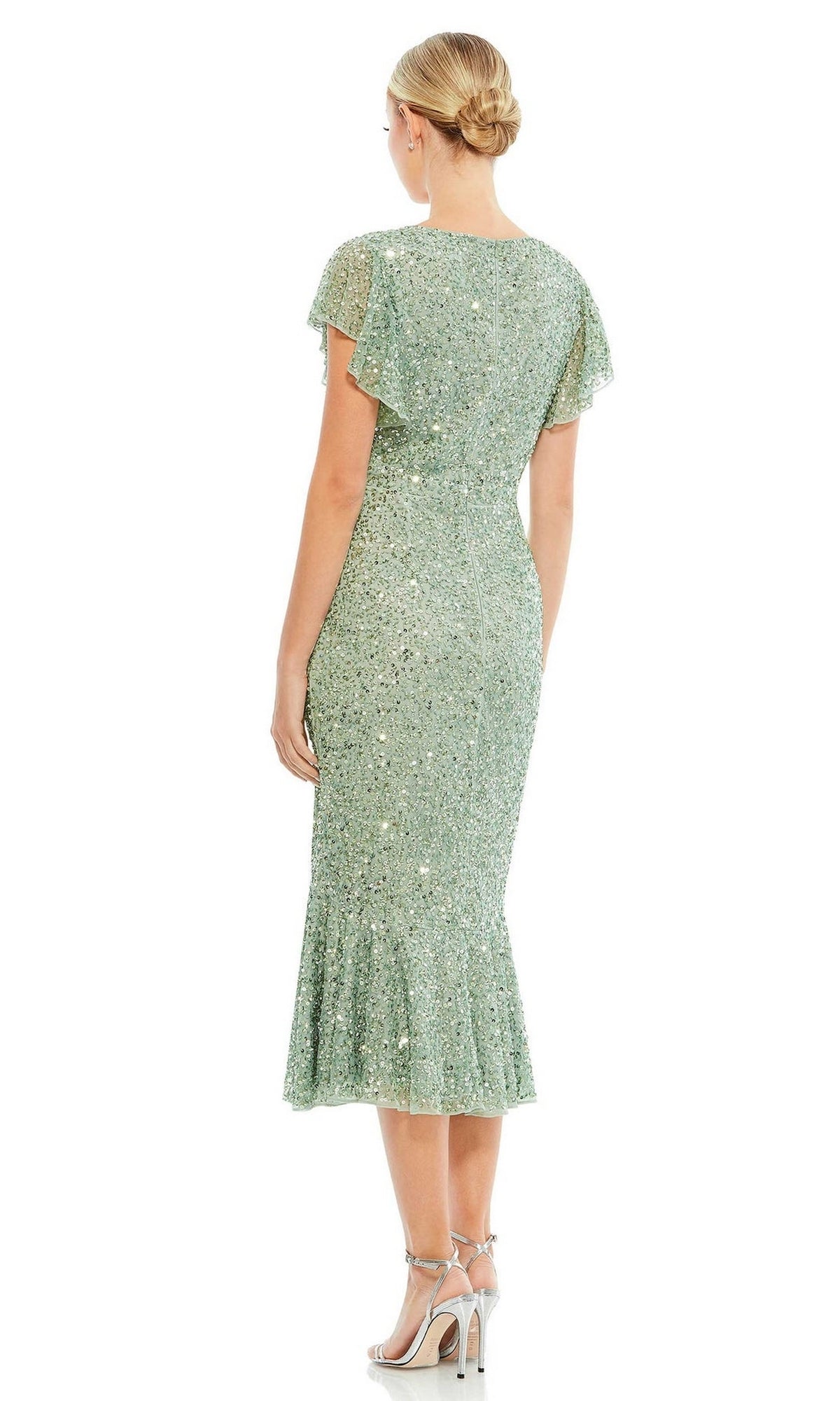 A woman stands facing away, wearing the Short Wedding Guest Dress 10801 by Mac Duggal in sage green sequins with short flutter sleeves and a ruffled hem, paired with strappy silver heels.