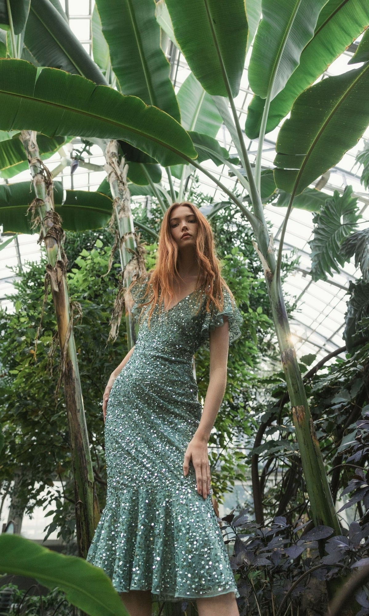 A woman in the Short Wedding Guest Dress 10801 by Mac Duggal, a sage green sequin dress, stands among large tropical plants inside a greenhouse.