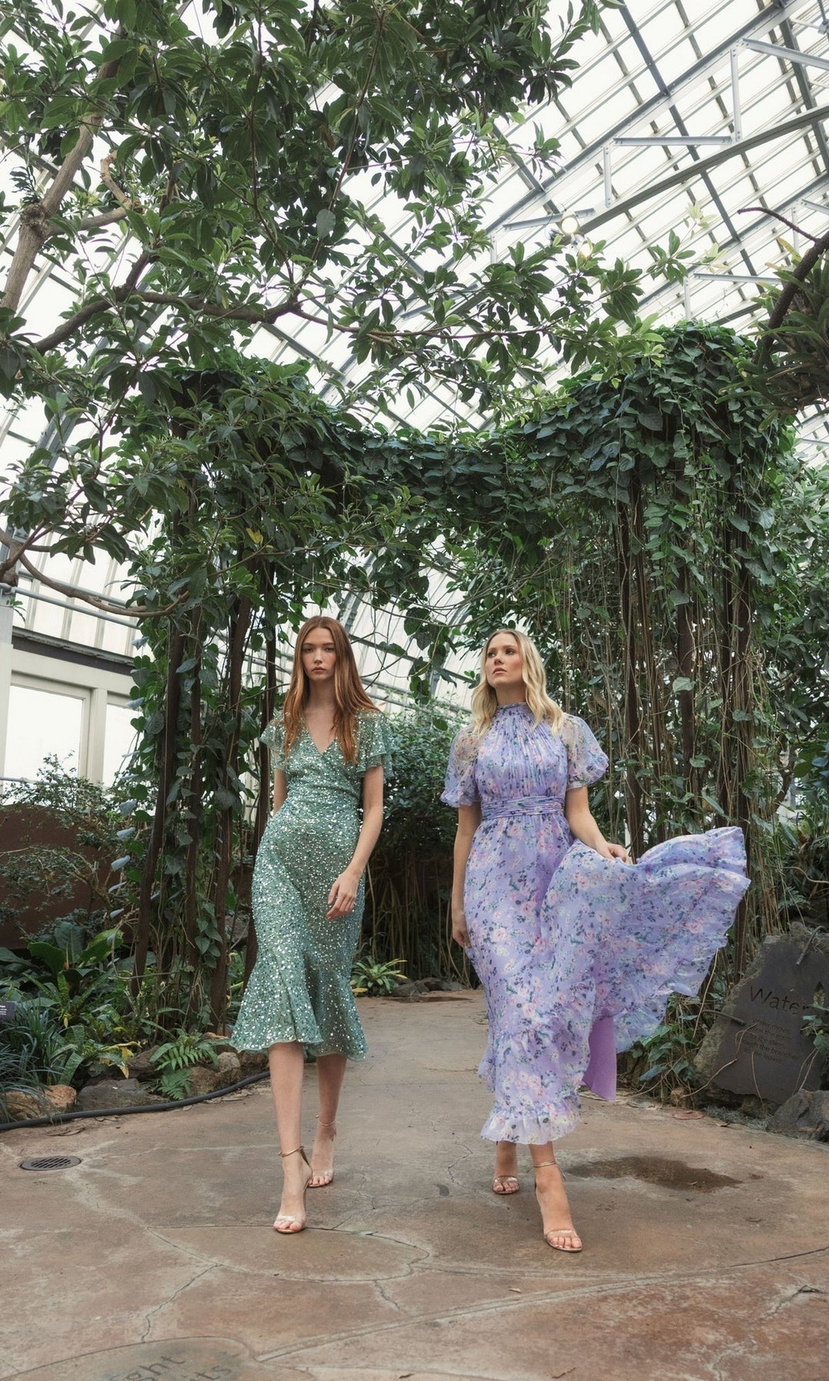 Two women wearing flowy, floral print dresses and one in the Mac Duggal Short Wedding Guest Dress 10801 walk along a garden path inside a greenhouse with lush greenery and a glass ceiling.