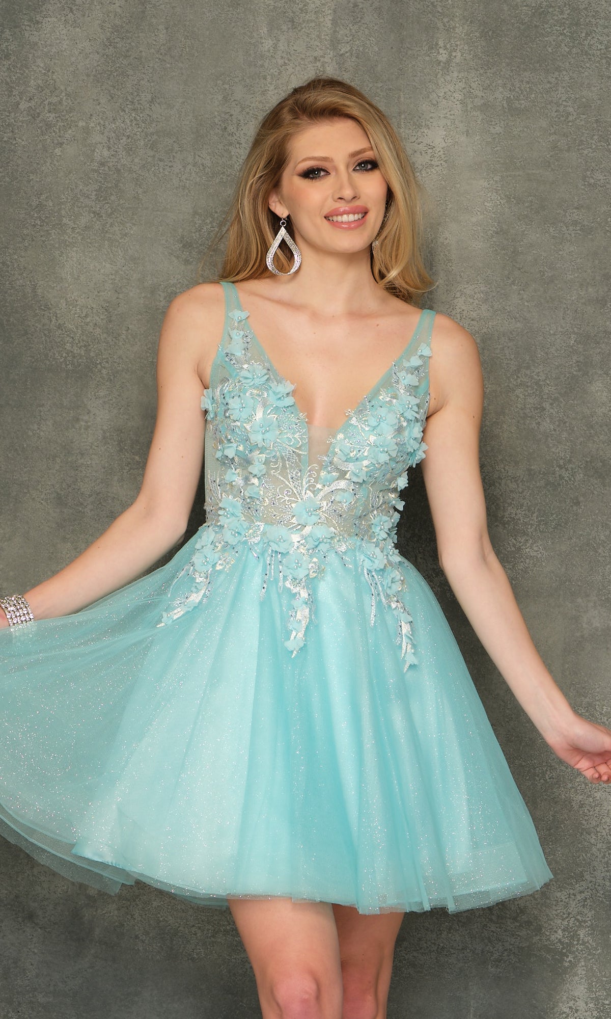 A woman wears the Sheer-Bodice Short Homecoming Dress—a sleeveless, light blue A-line tulle dress with floral accents—standing against a textured gray background and smiling at the camera.
