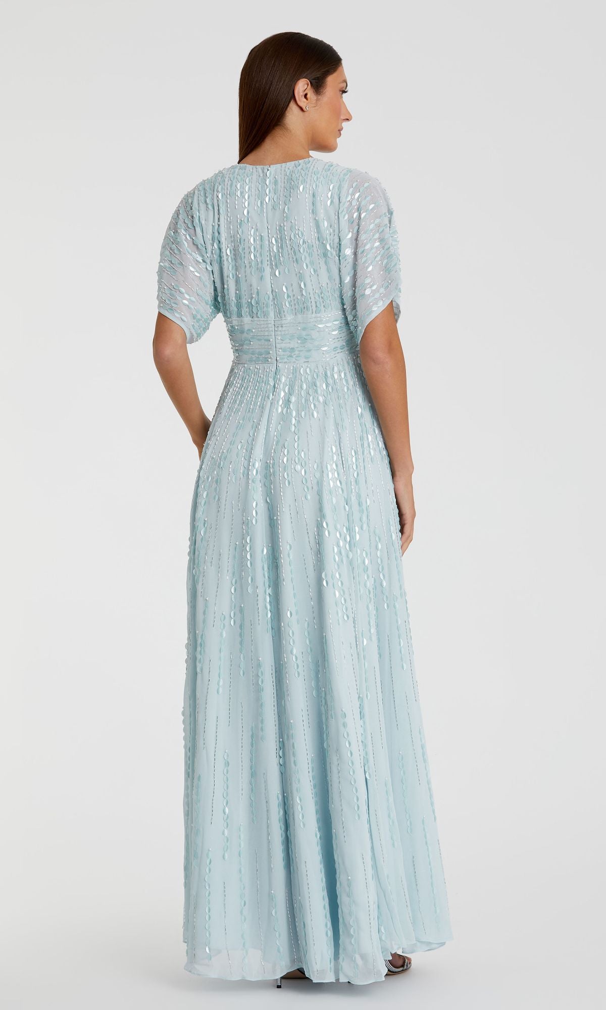 A woman stands facing away, wearing the Mac Duggal 10807 short sleeve blue formal dress—an elegant, modest a-line gown with shimmering sequin details.