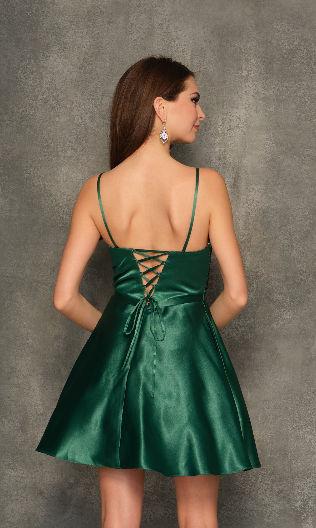 A woman faces away against a textured gray background, wearing the Beaded-Bodice Emerald Green Homecoming Dress 10845 featuring a short length, beaded bodice, and lace-up back.