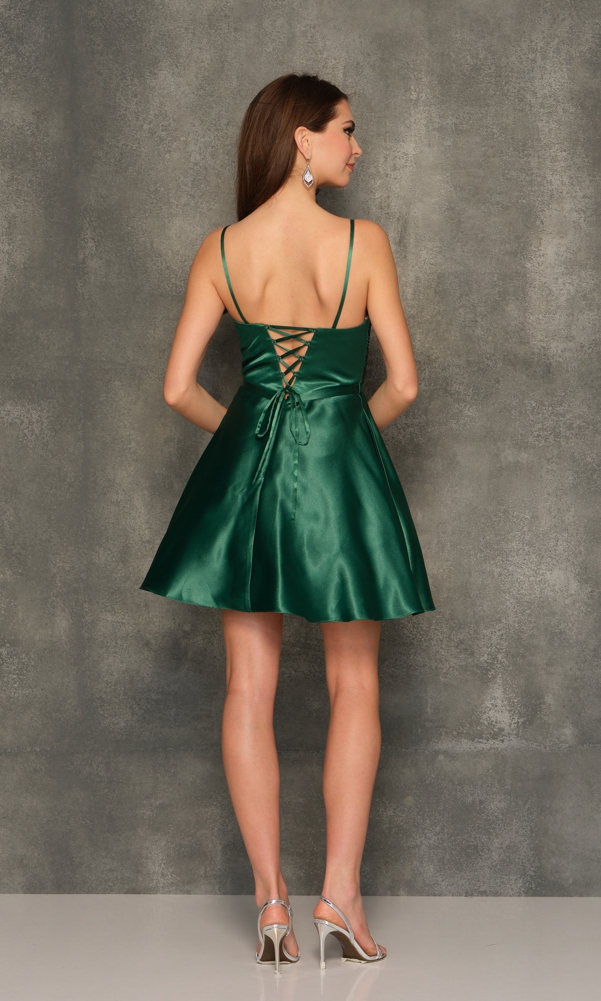 A woman models the Beaded-Bodice Emerald Green Homecoming Dress 10845, featuring thin straps, a lace-up back, and a shimmering satin A-line skirt, against a textured gray background.