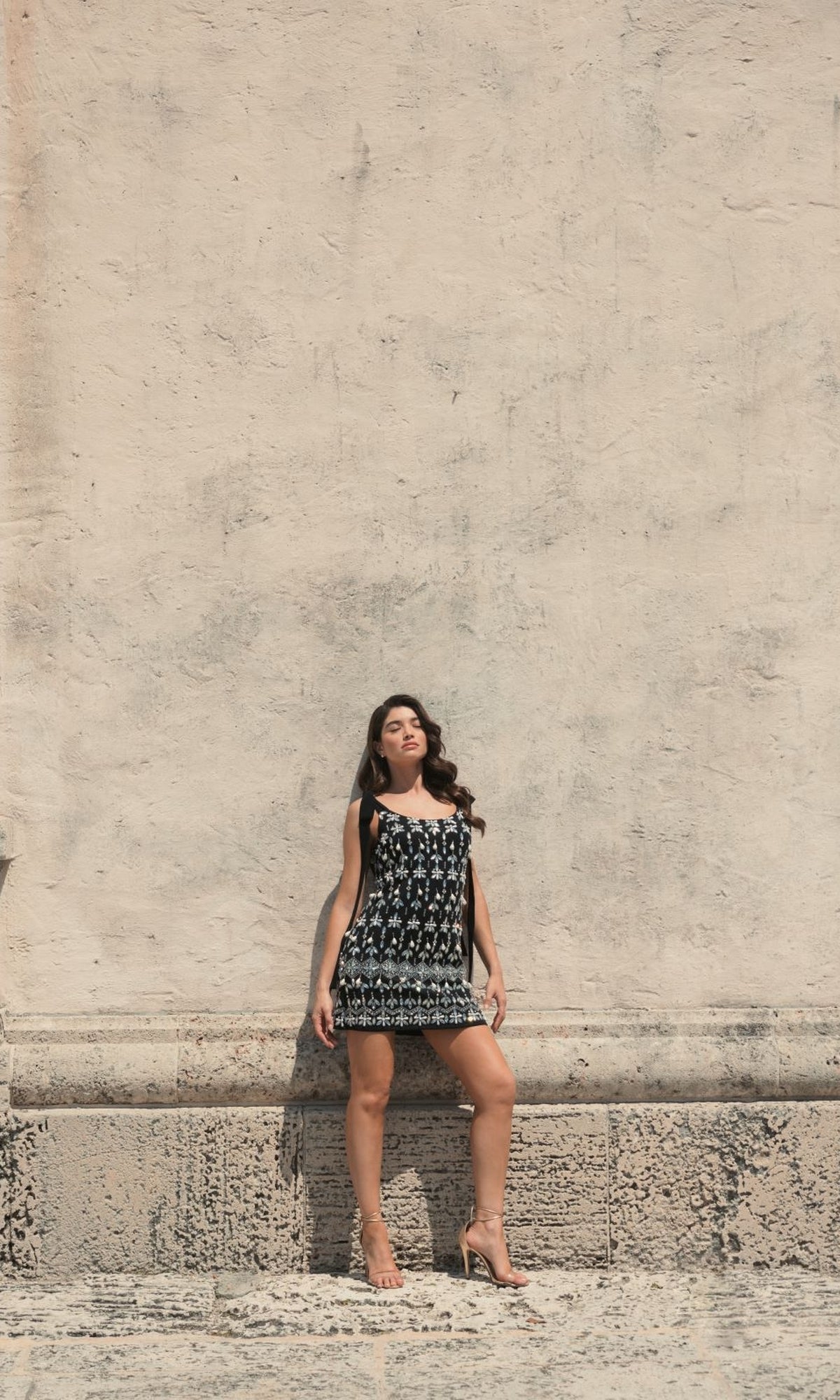 A woman in the Short Wedding Guest Dress 10937 by Mac Duggal stands against a textured beige wall, eyes closed and hands by her sides, exuding relaxed elegance.