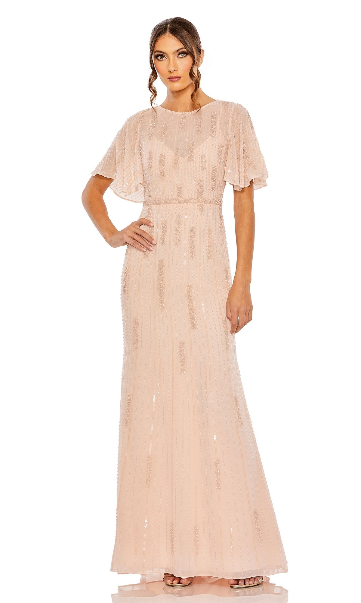 A woman wears the Long Formal Dress 11014 by Mac Duggal, a floor-length blush pink gown with a v-neckline, sheer short sleeves, and sequin accents, posed against a plain white background.