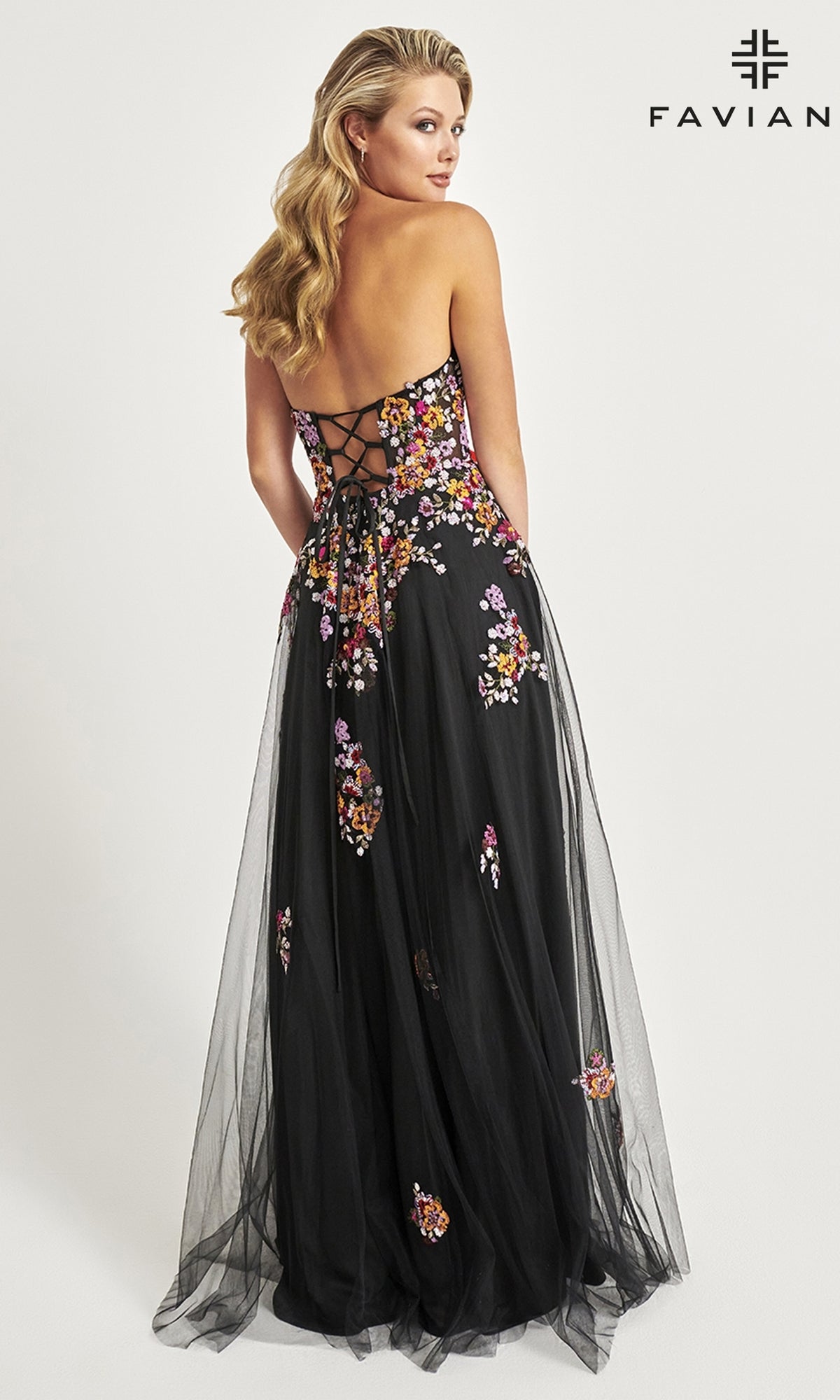A woman faces away in the Faviana Long Black Strapless Prom Dress 11028, a floor-length backless gown with floral embroidery and lace-up detailing.