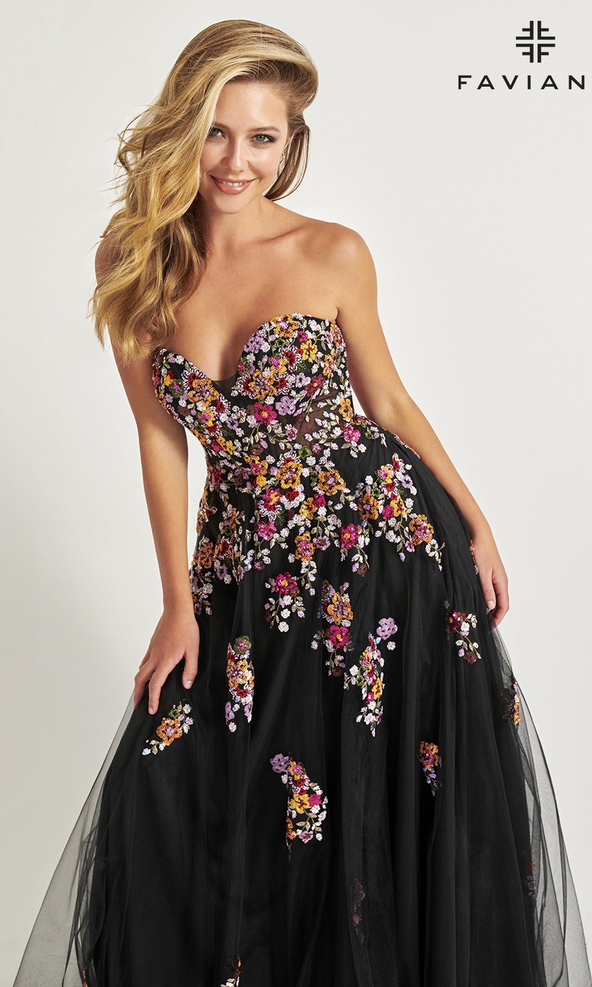 A woman models the Faviana Long Black Strapless Prom Dress 11028, featuring colorful floral embroidery, against a plain backdrop with the Faviana logo in the top right corner.