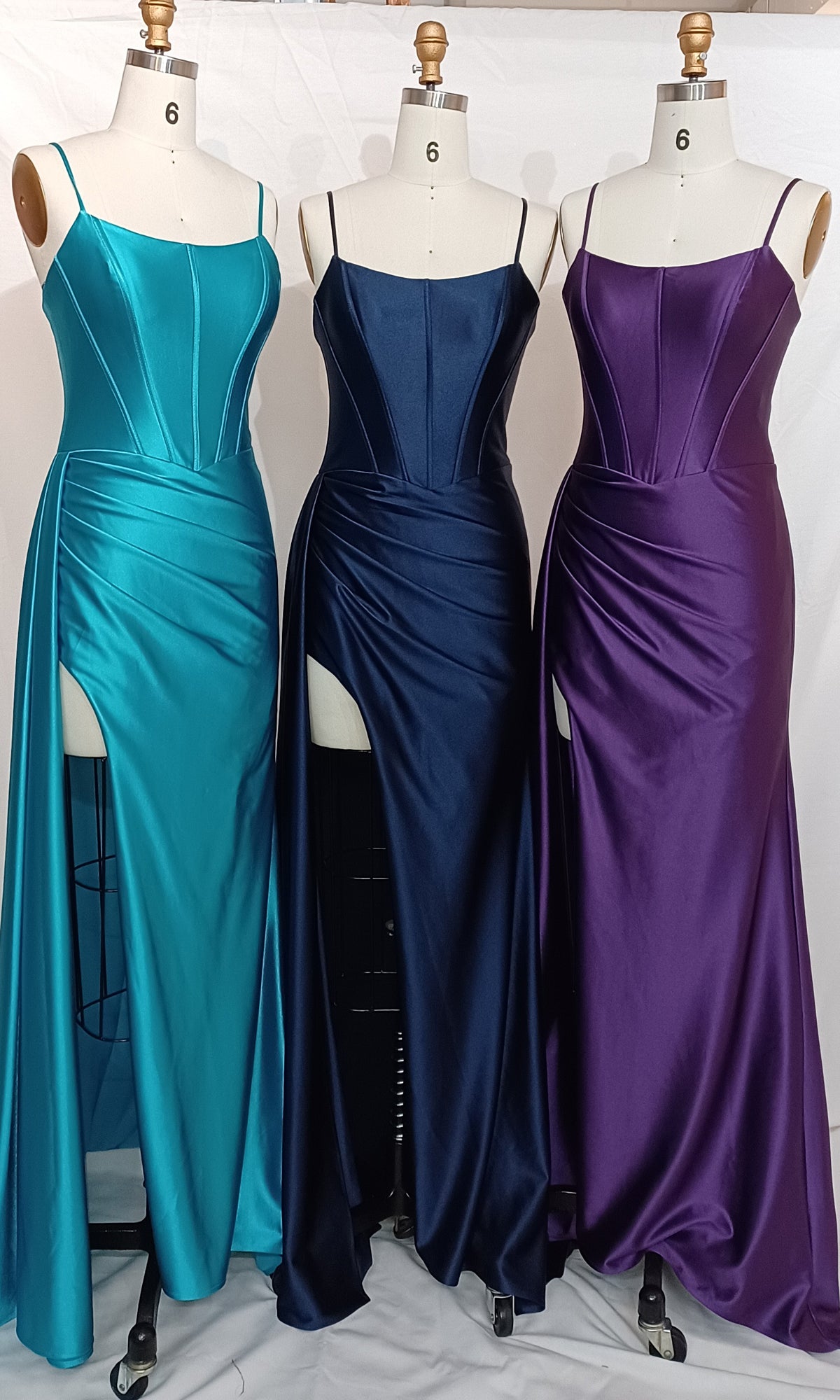 Three mannequins display Dave & Johnny Long Satin Prom Dresses with side sash in teal, navy blue, and purple, each featuring thin straps, high slits, and a fitted corset-style bodice, arranged side by side.