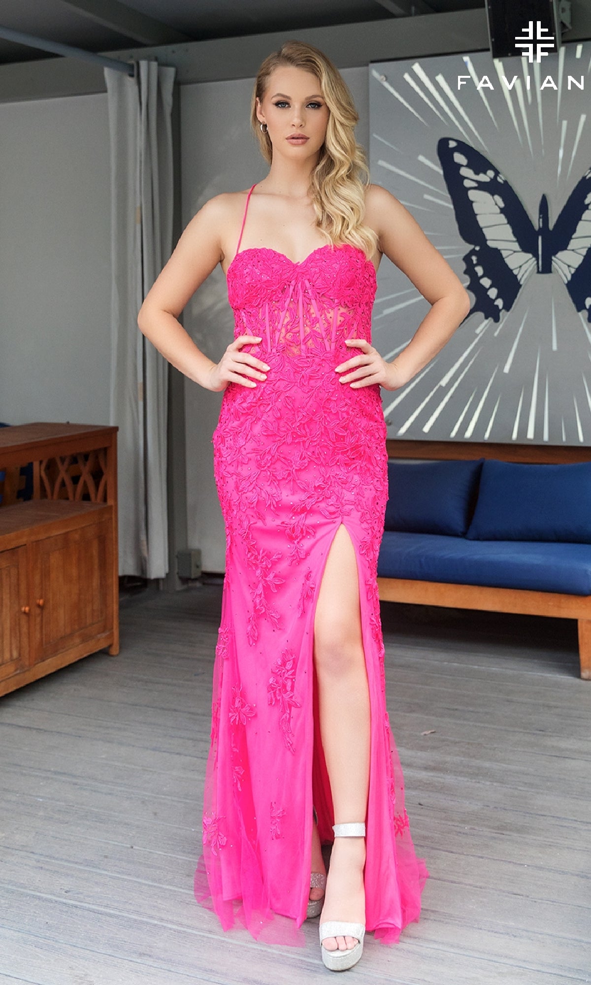 A woman wears the Lace-Applique Long Faviana Prom Dress 11054—a bright pink, strapless gown with lace appliques and a thigh-high slit—standing indoors by a blue sofa and butterfly wall art.