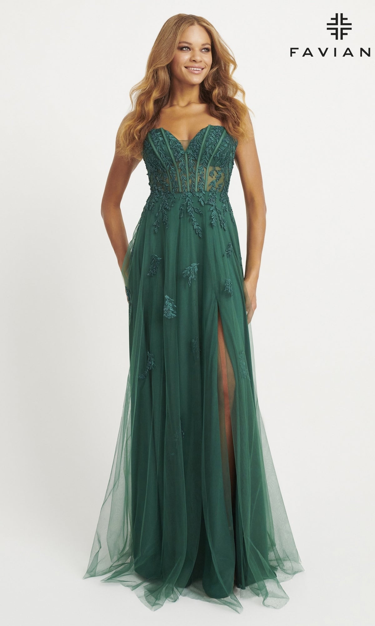 A woman models the Strapless Tulle Faviana Long Prom Dress 11057 in deep green, featuring a sheer corset bodice, floral appliqués, a high front slit, and a floor-length tulle skirt, standing against a plain white background.