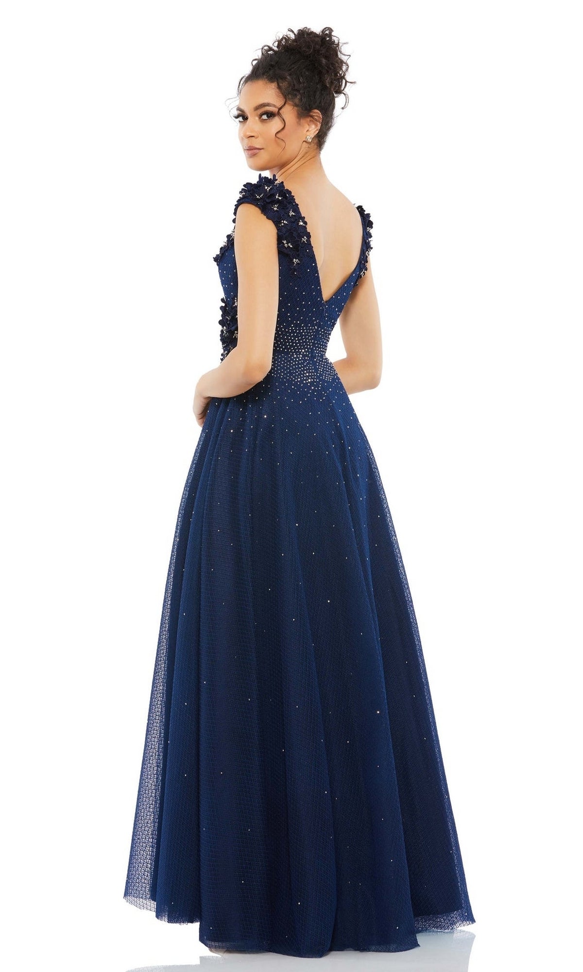 A woman wears the Mac Duggal V-Neck Midnight Blue Formal Dress 11152, featuring a floor-length silhouette, deep V-back, and embroidered floral appliques, as she stands facing away and looks over her shoulder.