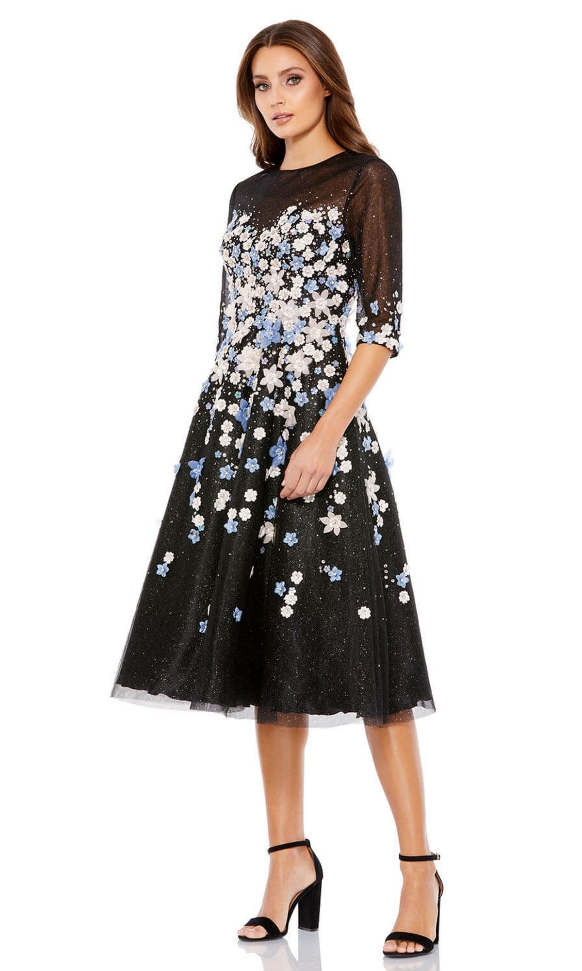 A woman wearing the Short Wedding Guest Dress 11161 by Mac Duggal—black, knee-length with floral embroidery and sheer sleeves—pairs it with black high-heeled sandals, standing against a white background.