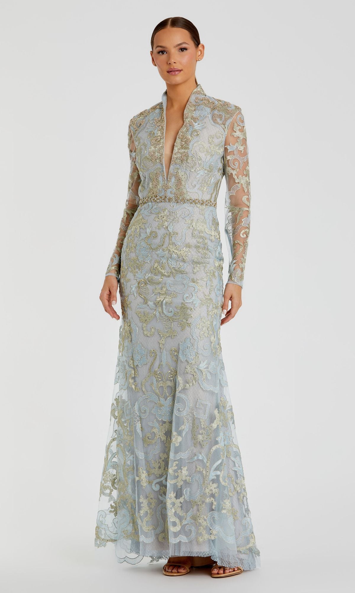 A woman models the Mac Duggal 11165 Long Sleeve Lace Formal Gown, featuring a light blue vintage design with gold embroidery, deep V neckline, long sleeves, and floor-length silhouette against a plain background.