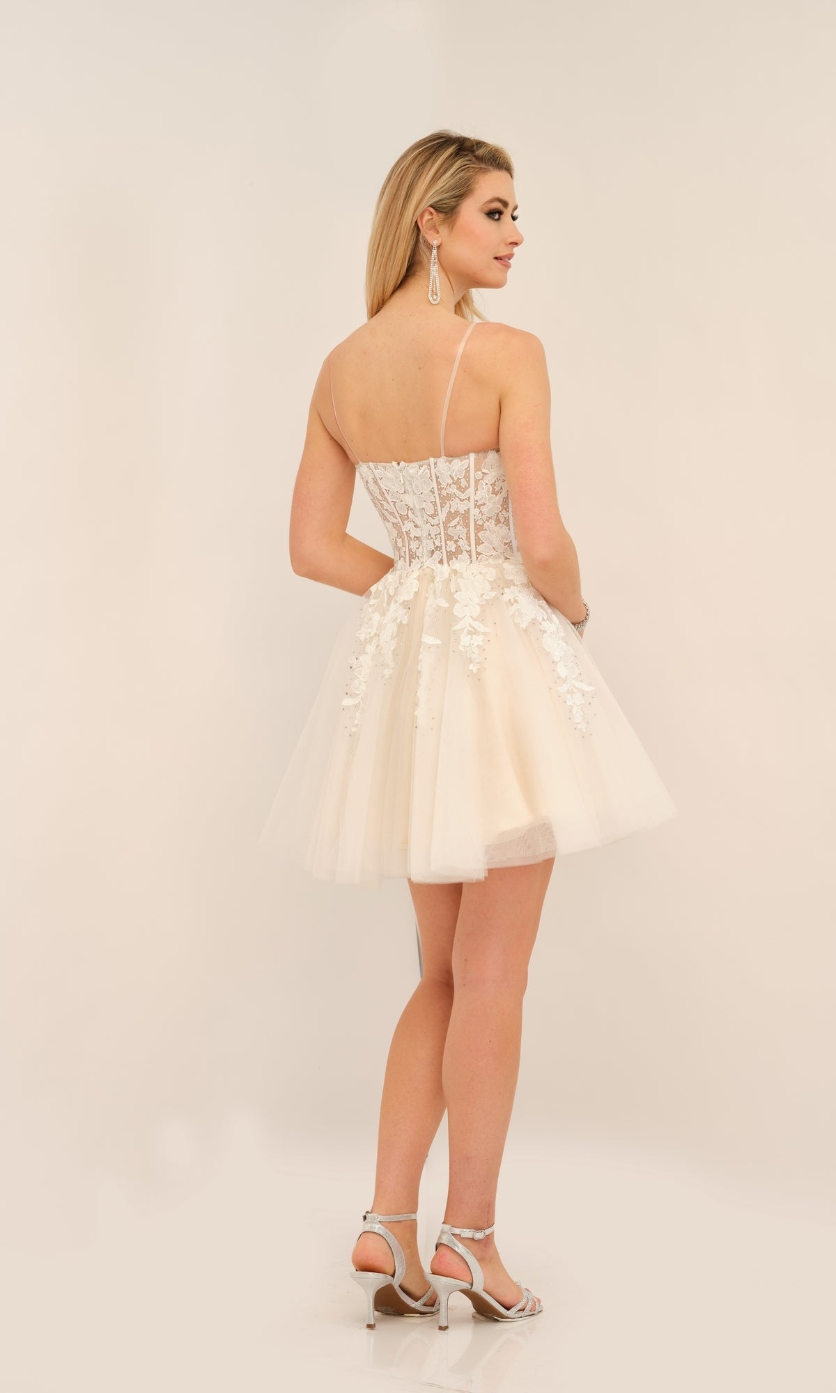 Woman wearing the Dave and Johnny Short White Hoco Dress 11206 with a sheer corset bodice, thin straps, and white high-heeled sandals—an embroidered lace party dress ideal for special occasions.