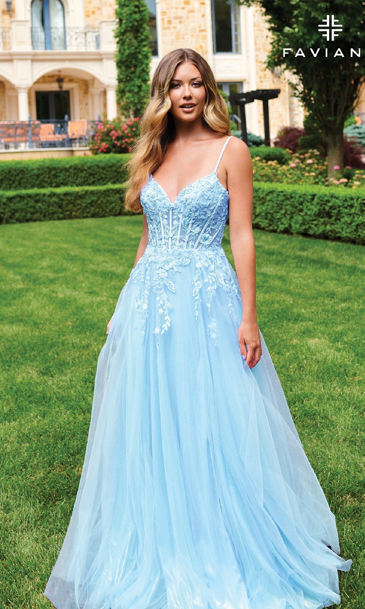 A woman stands on a manicured lawn before an elegant building, wearing the Faviana 11230—a long, light blue A-line prom dress with a lace bodice, fitted corset top, tulle skirt, and delicate floral applique.
