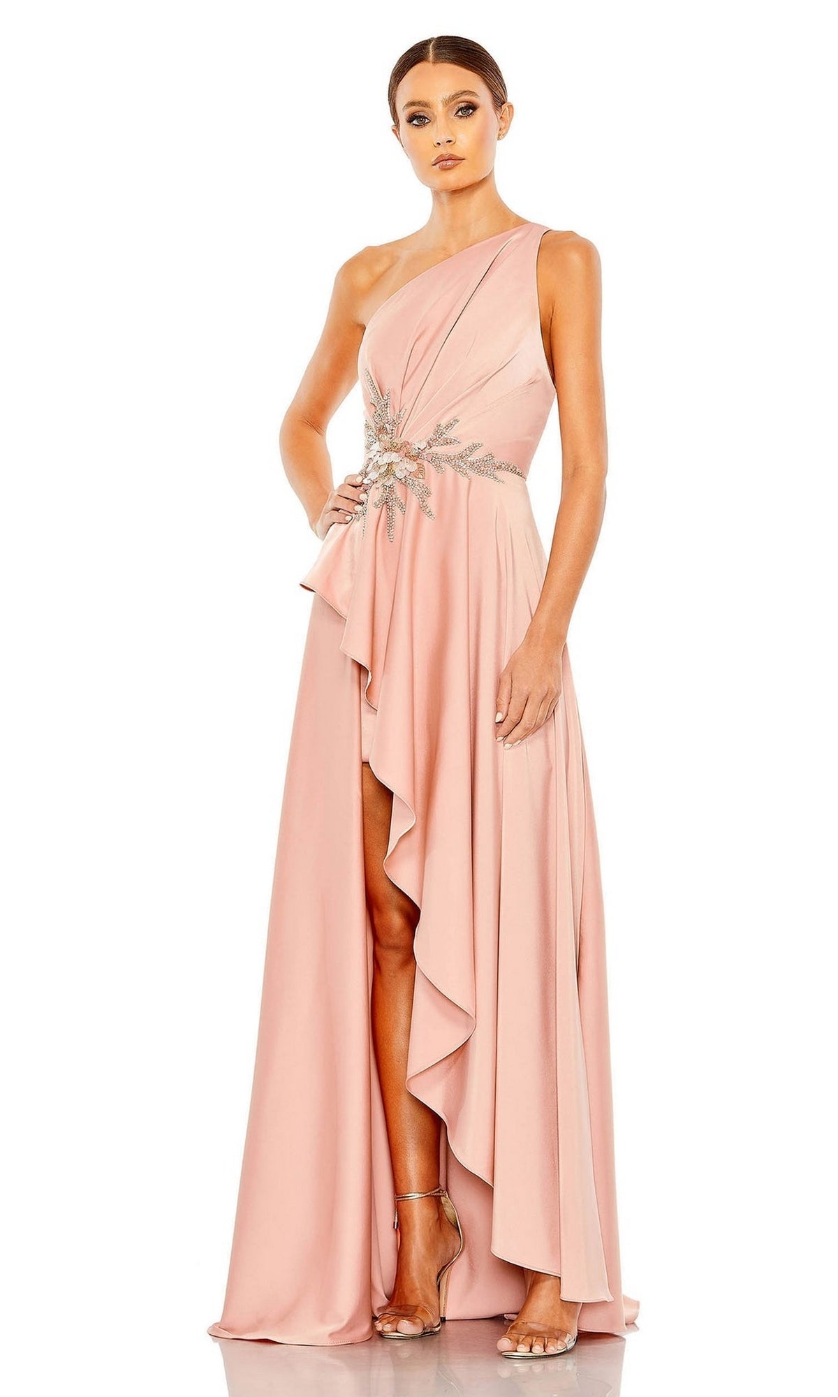 A woman stands elegantly in the Mac Duggal Long Formal Dress 11262, a blush pink one-shoulder gown featuring an embellished waist and asymmetrical ruffled hem, paired with strappy heels.