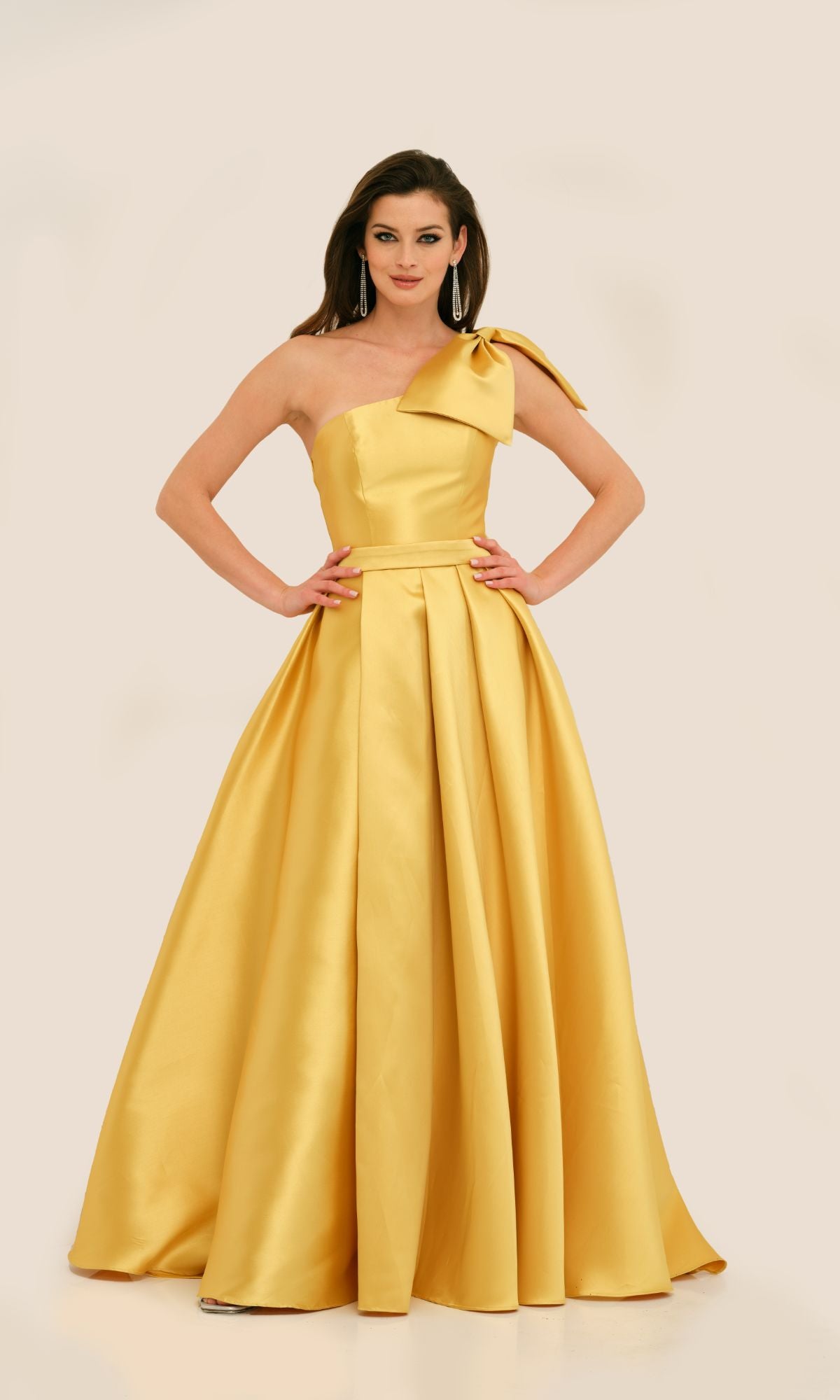 A woman stands with hands on hips against a plain background, wearing the Long Formal Dress 11337 by Dave and Johnny—a floor-length, golden yellow one-shoulder ball gown featuring a bow detail.