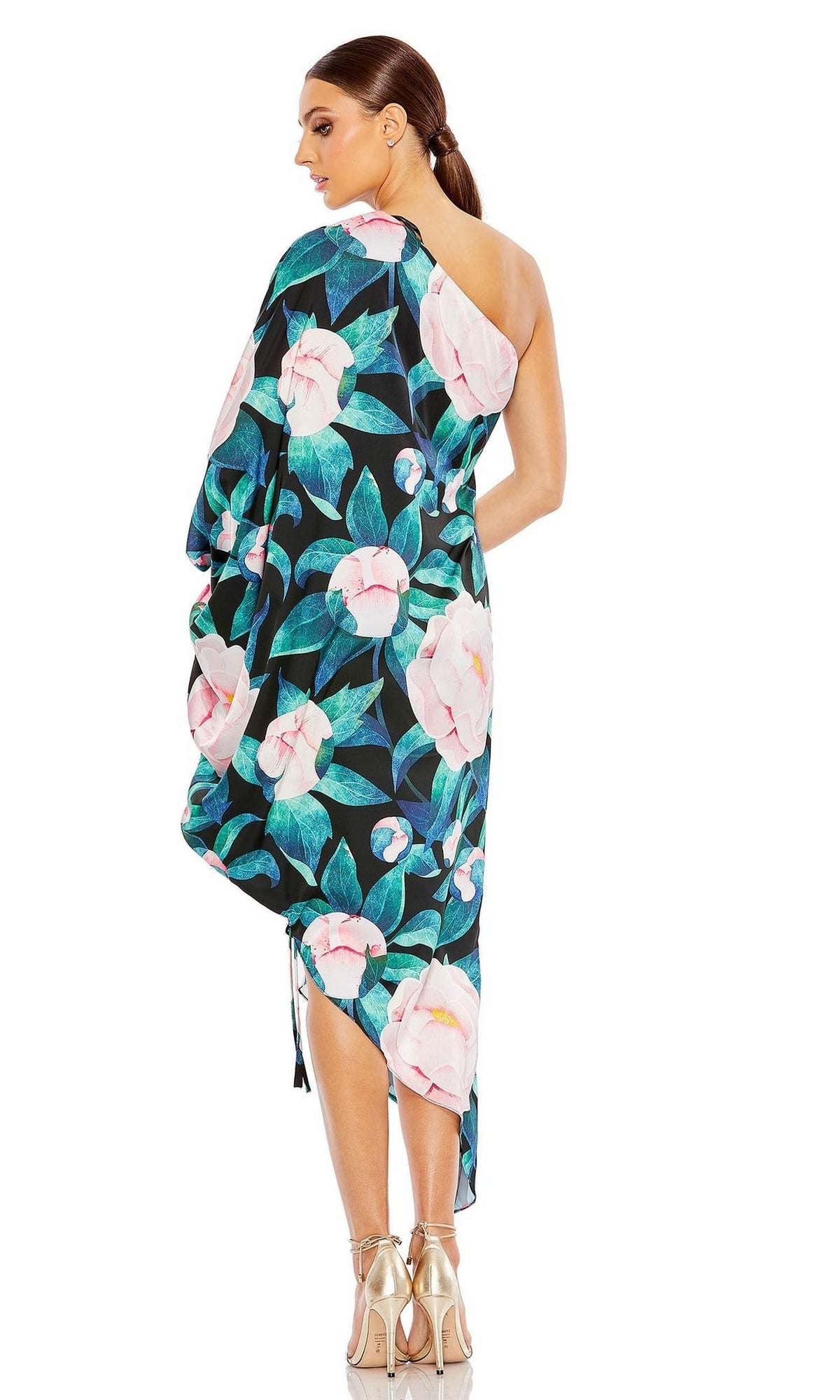 A woman with brown hair in a ponytail wears the Mac Duggal 11404 Short Print Wedding Guest Dress, featuring a one-shoulder high-low floral design, and pairs it with gold high heels while standing with her back to the camera.
