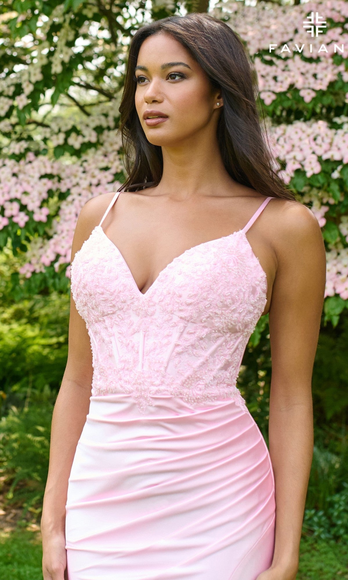 A woman wearing the Faviana 11406 long prom dress with a light pink satin skirt and lace-accented bodice stands outdoors amid blooming pink flowers and greenery.