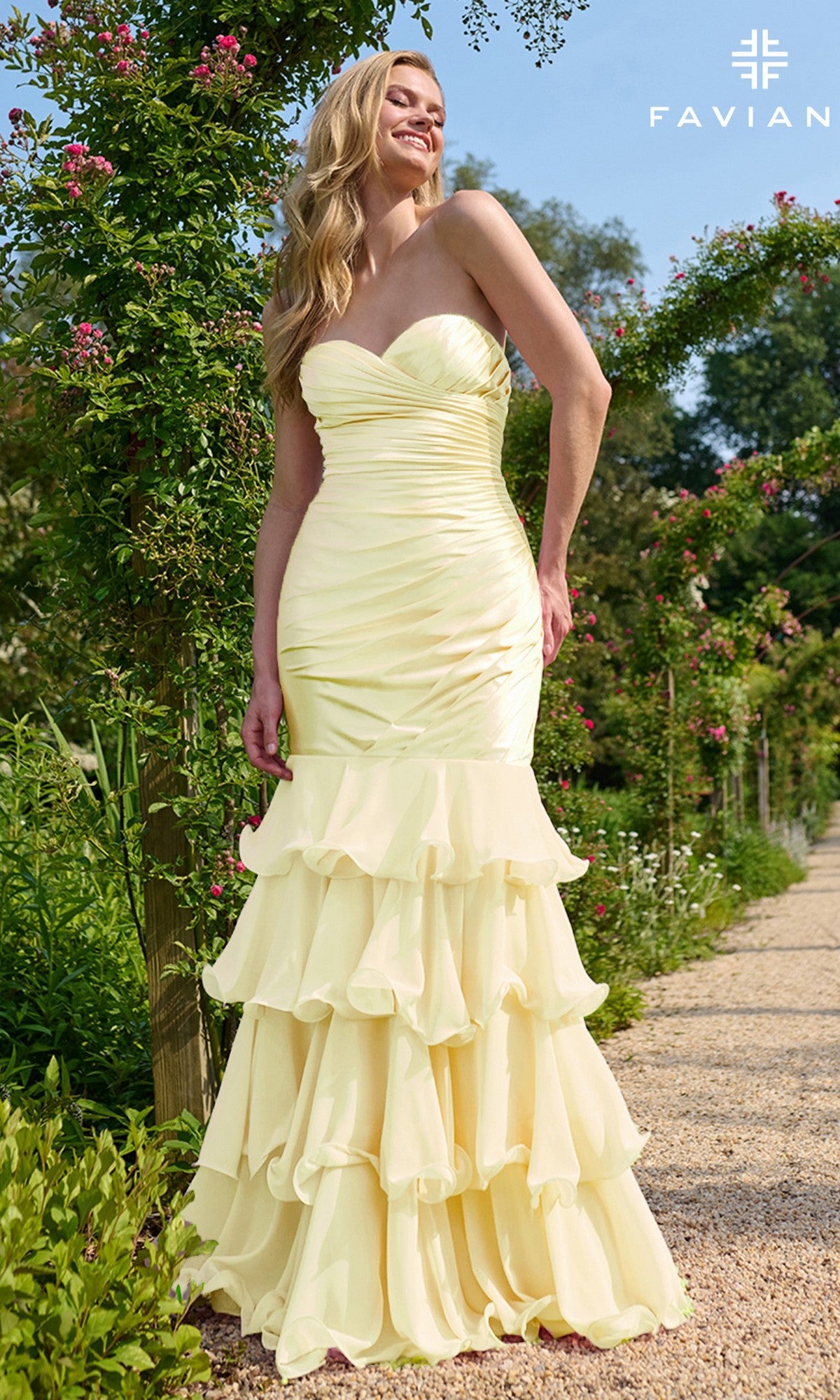 Wearing the Faviana 11419 long prom dress, a woman stands on a garden path, her ruffled pastel yellow gown with a strapless sweetheart neckline flowing amid lush greenery and blooming flowers.