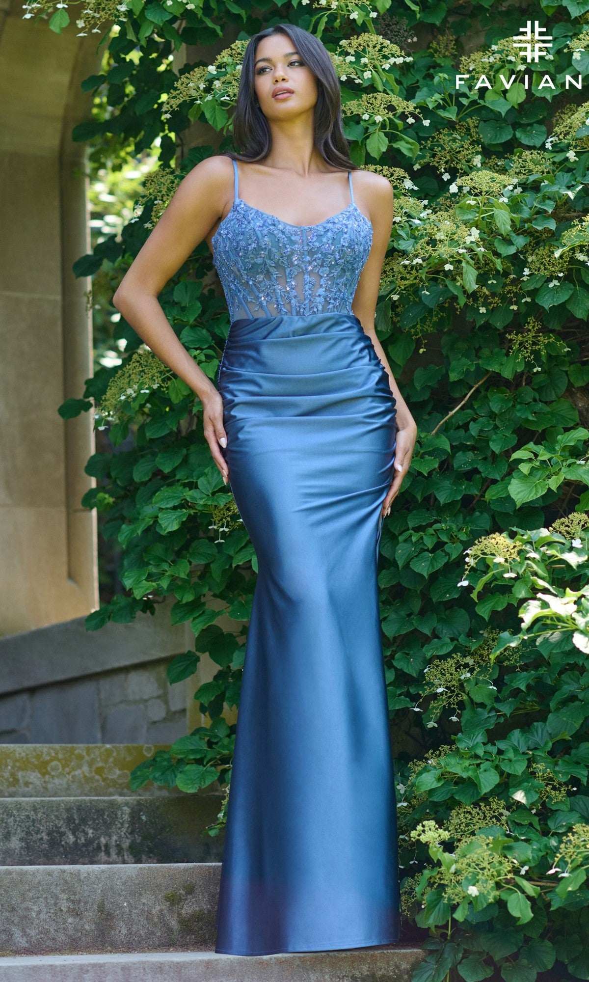 A woman wearing the Faviana 11433 Sheer Lace Corset Long Prom Dress stands on stone steps before green foliage. The dress is blue, fitted, sleeveless, with a beaded corset-style bodice.