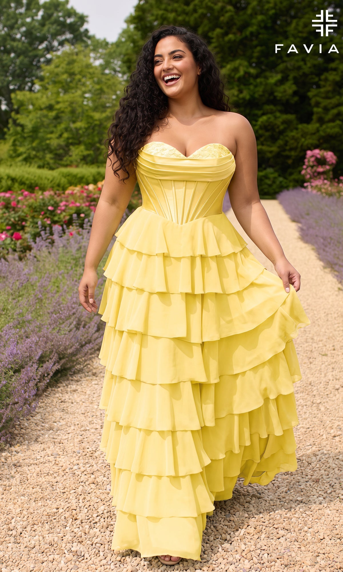 A woman walks along a gravel garden path wearing a Faviana 11436E Long Plus-Size Prom Dress, a light yellow strapless gown featuring a corset bodice and a tiered ruffled chiffon skirt.
