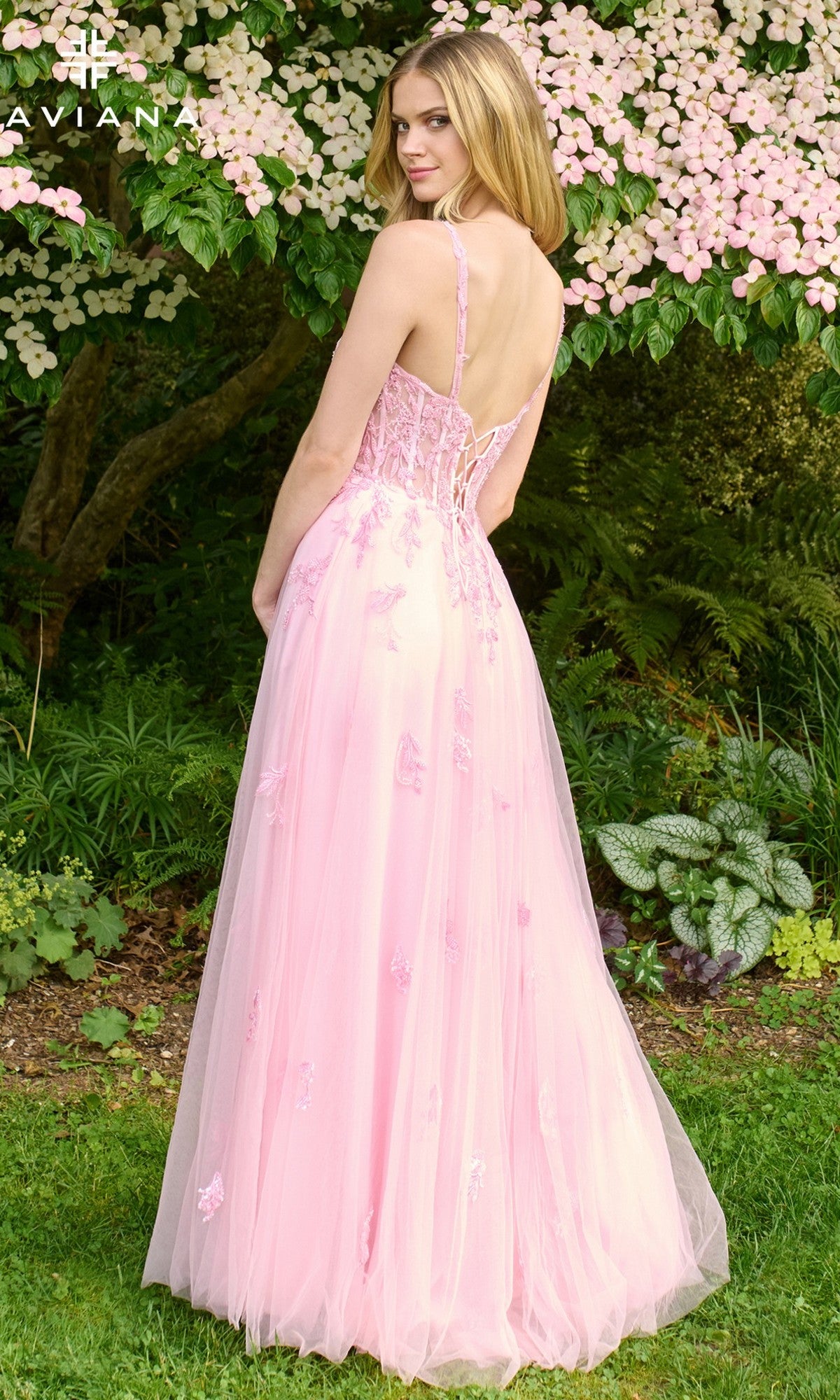 A woman wearing the Faviana 11437 long prom dress in light pink with lace appliqué stands outdoors before green foliage and pink flowers, glancing over her shoulder.