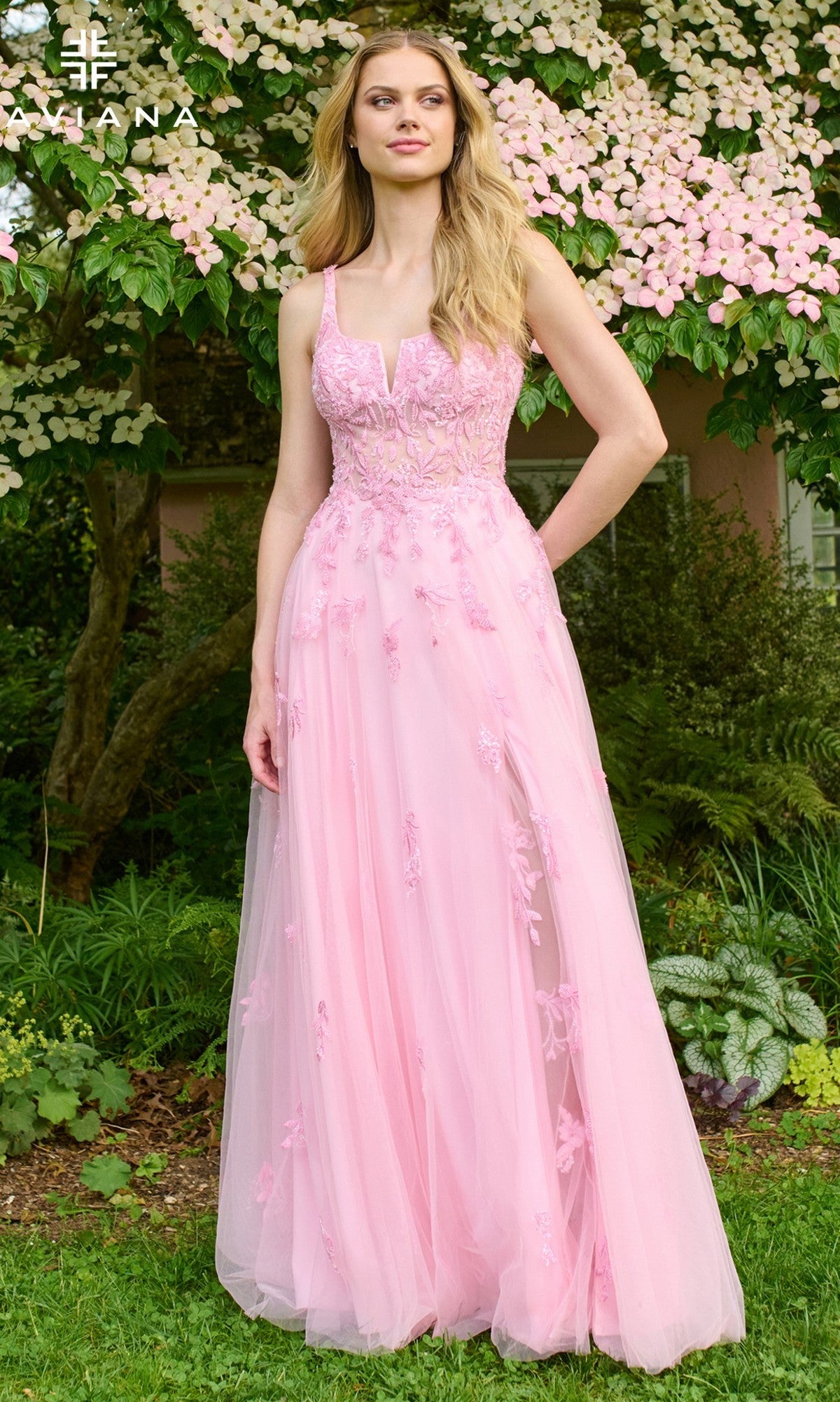 A woman stands outdoors in the Faviana 11437 long prom dress—a light pink, sleeveless, A-line gown with floral embroidery and a fitted bodice. Lush greenery and pink flowers in the background highlight her elegant tulle look.