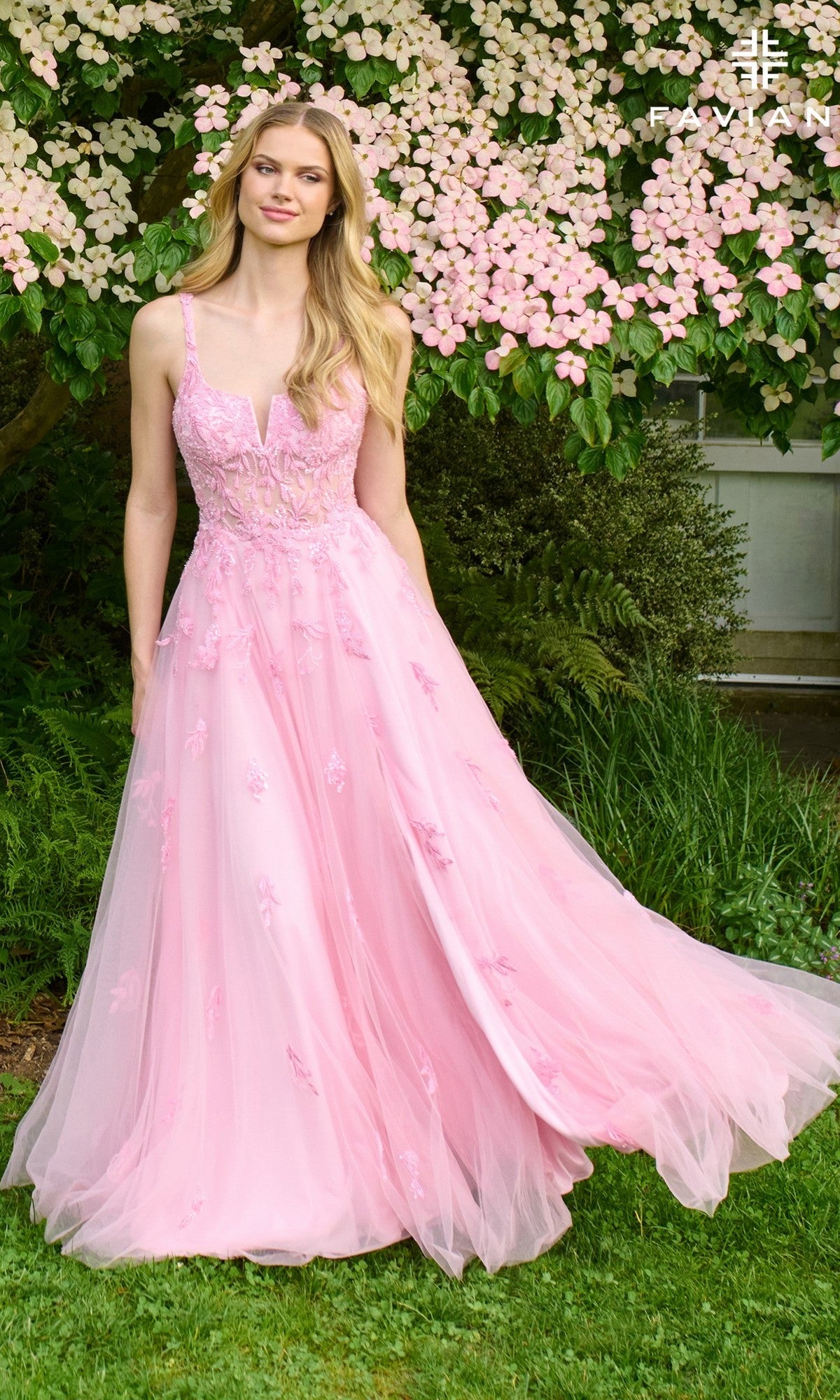 A woman in the Faviana 11437 long prom dress, featuring light pink tulle and delicate lace applique, stands before a blooming tree with pale pink flowers and lush greenery in the background.