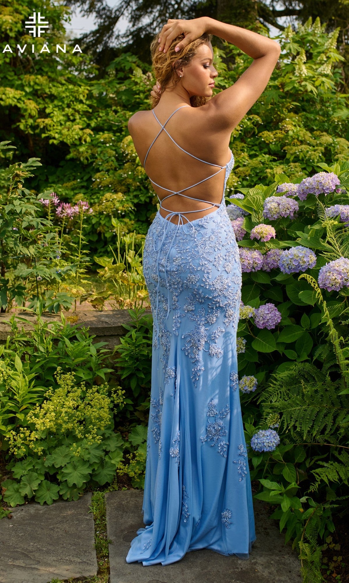 A woman stands in a garden wearing Faviana 11440, a light blue long prom dress with an open back, strappy details, and beaded applique, surrounded by green foliage and blooming hydrangeas.