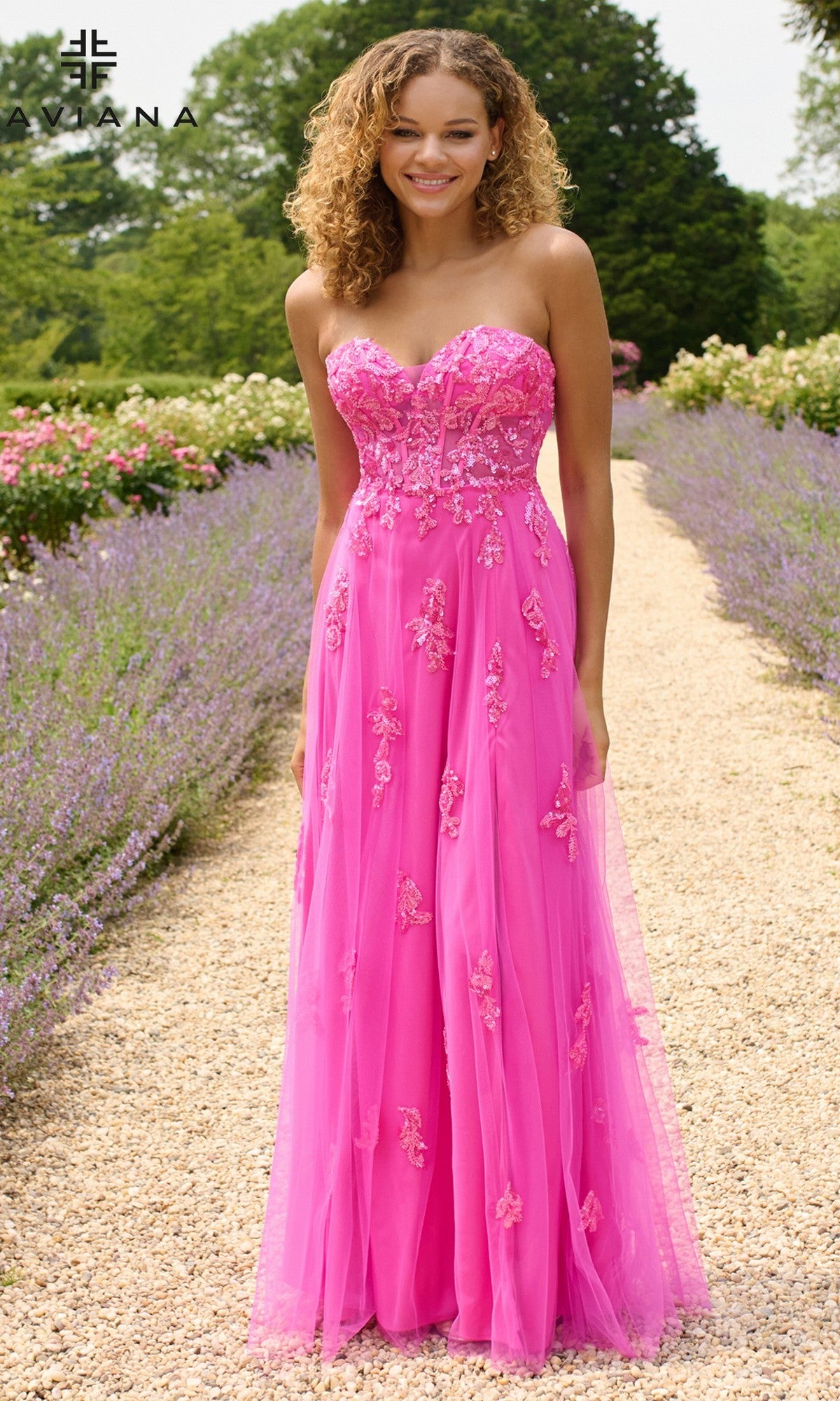 A woman wears the Faviana 11474 long prom dress, featuring a strapless corset bodice and bright pink floral embroidery, while standing on a garden path surrounded by green trees and purple flowers.