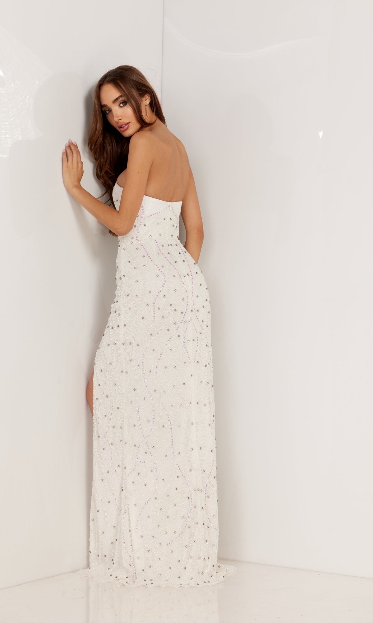 A woman in the Deep V-Neck Strapless Long Beaded Prom Dress 1148, adorned with sequins, stands against a white wall, facing away and looking over her shoulder.