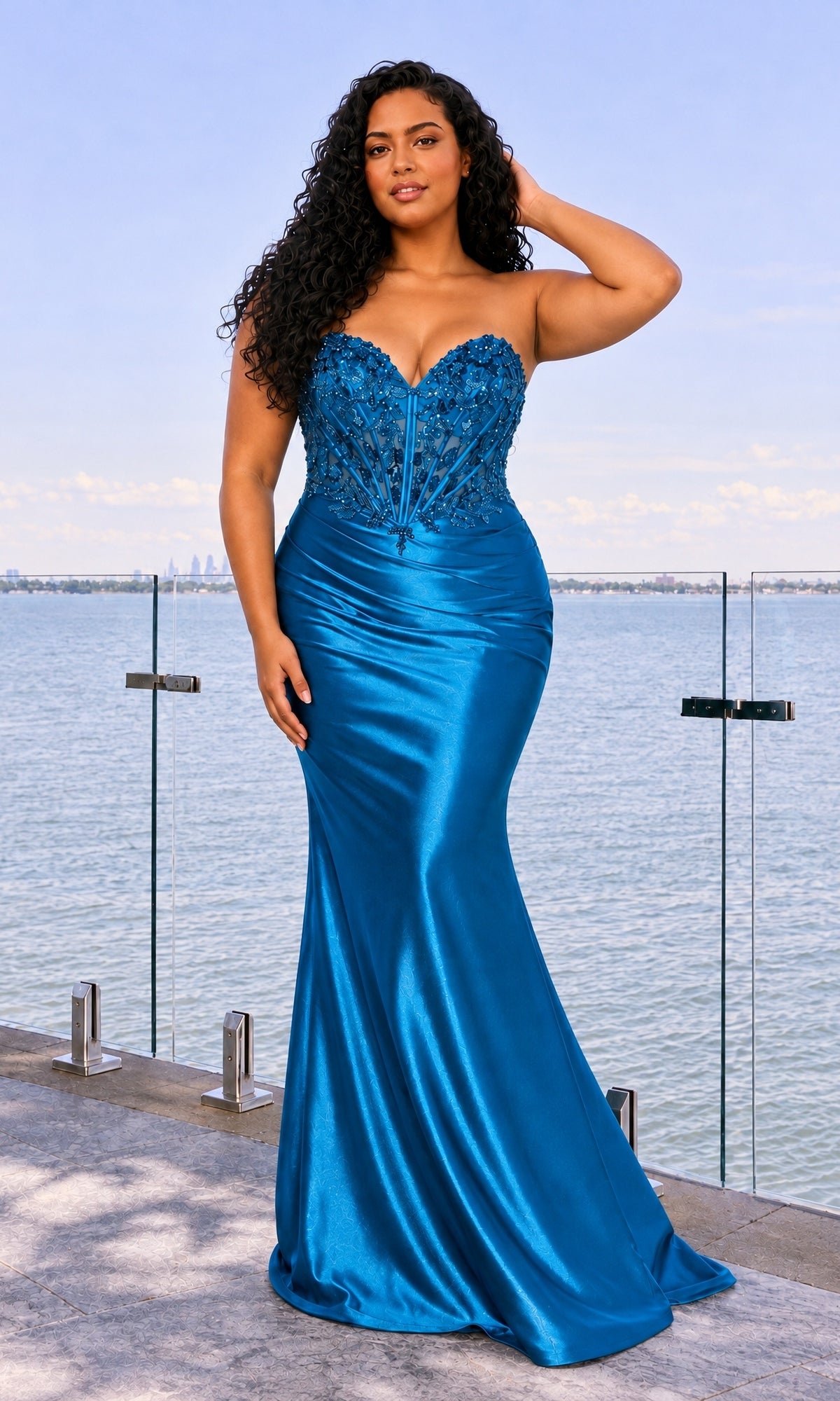 A person wears a strapless, ink blue, mermaid-style Faviana 11607E plus-size prom dress with a beaded corset bodice by a waterfront.