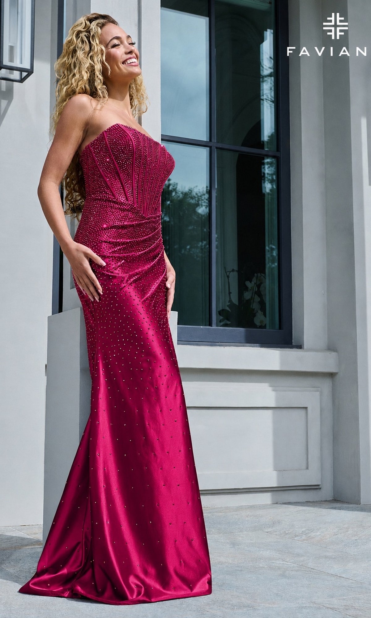 A woman wearing a cherry-colored Faviana 11613 long prom dress, a strapless, floor-length corset gown with scattered rhinestone embellishments.