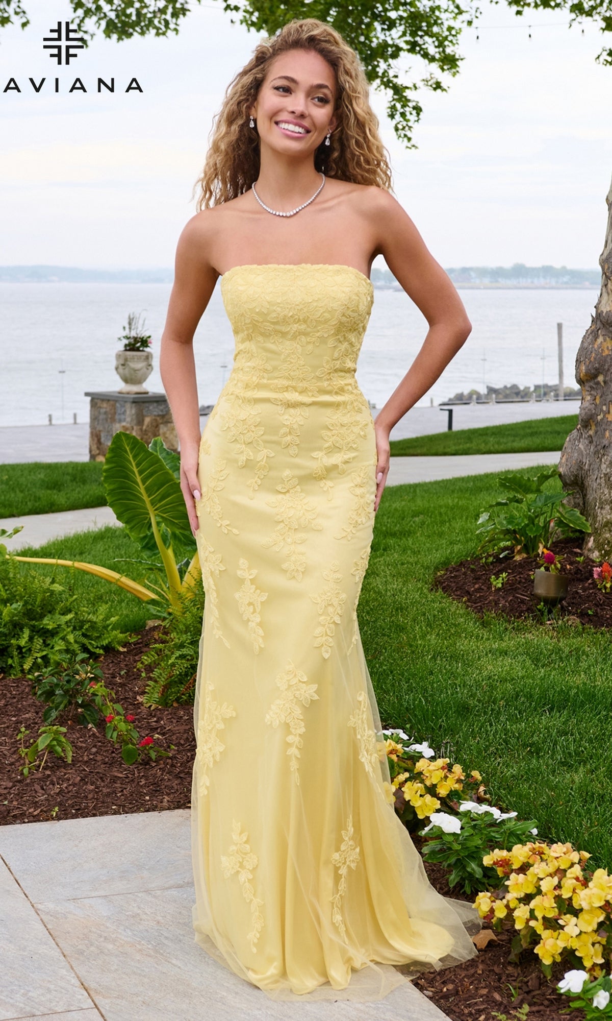 A woman wears a light yellow Faviana 11627 strapless column prom dress featuring a floor-length silhouette with delicate floral lace overlays.
