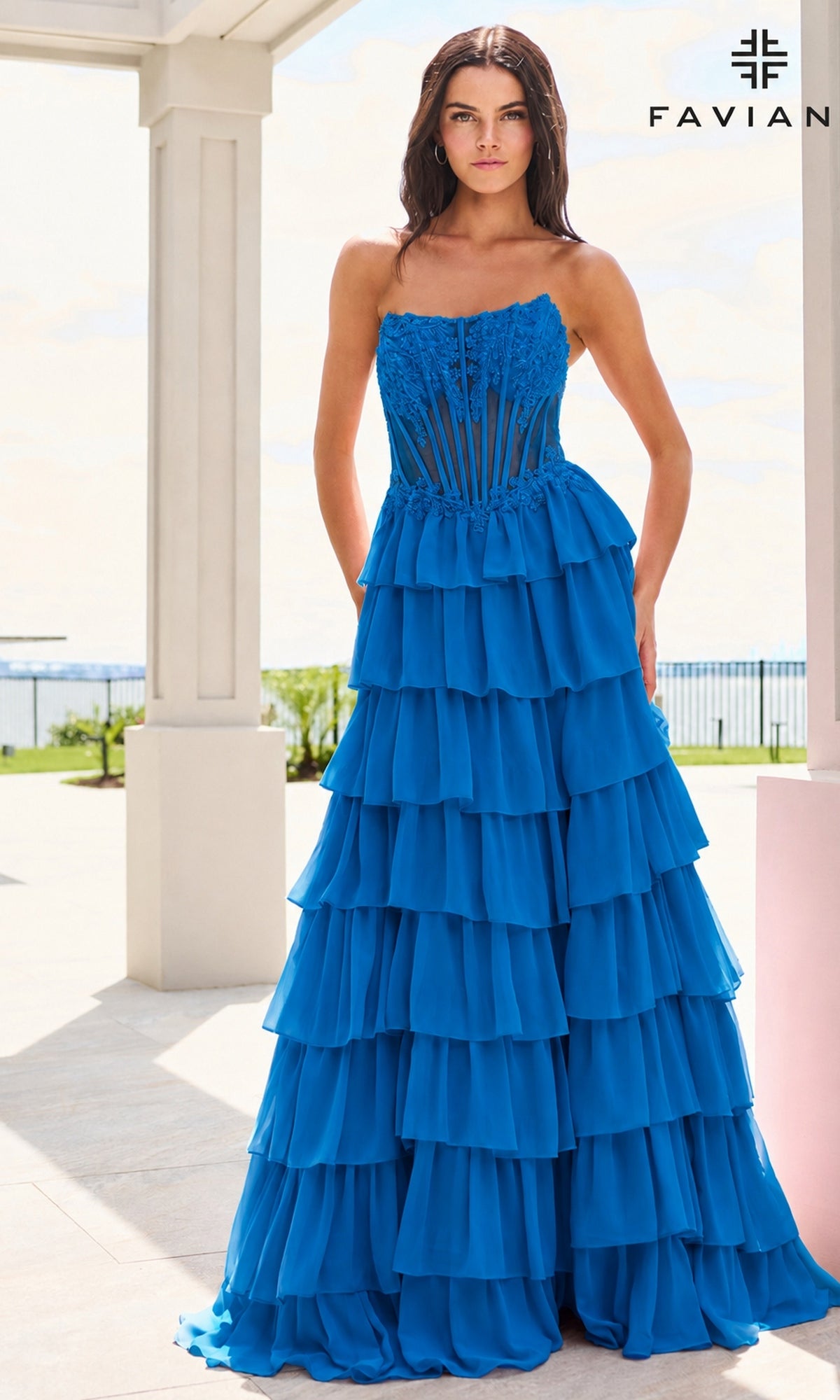 A model wearing a Faviana 11629 strapless, ink blue floor-length gown with a corset bodice and a tiered chiffon ruffle skirt.