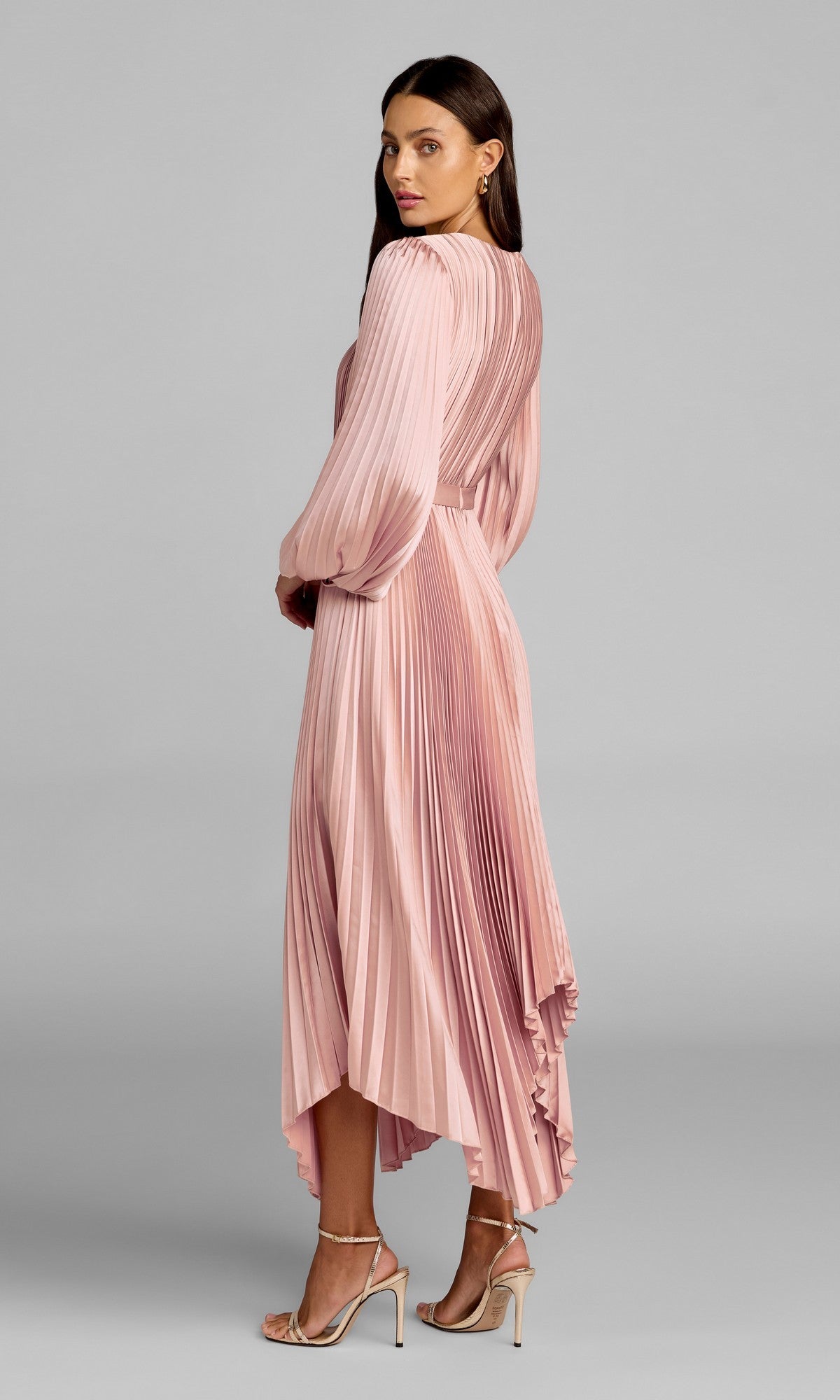 Woman in profile wears a fuchsia Mac Duggal 11638 midi wedding guest dress with pleats, long sleeves, and an asymmetrical hem, looking over her shoulder.