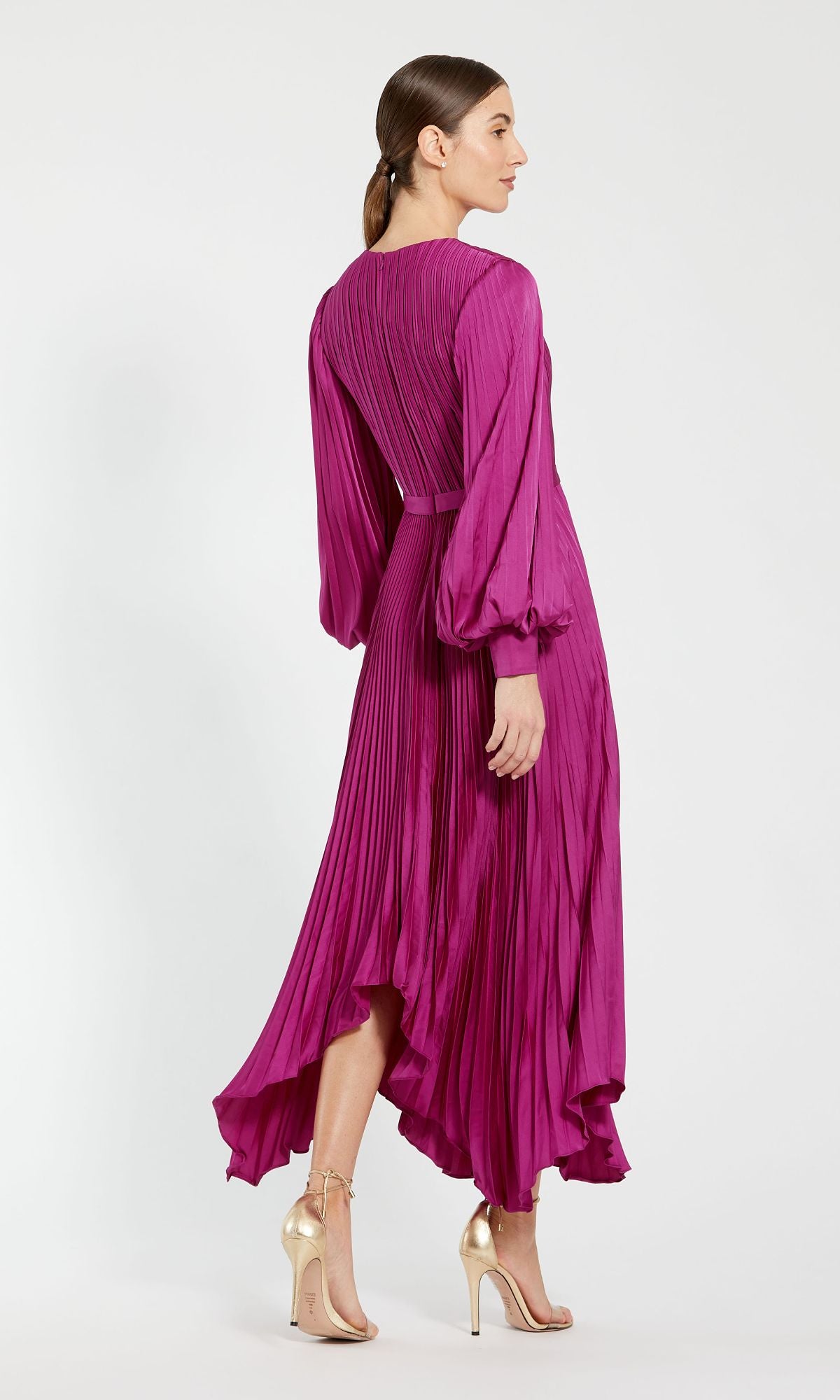 A woman faces away in the Mac Duggal 11638 Fuchsia Midi Wedding Guest Dress with pleats and long sleeves, paired with gold high-heeled sandals, set against a plain light background.