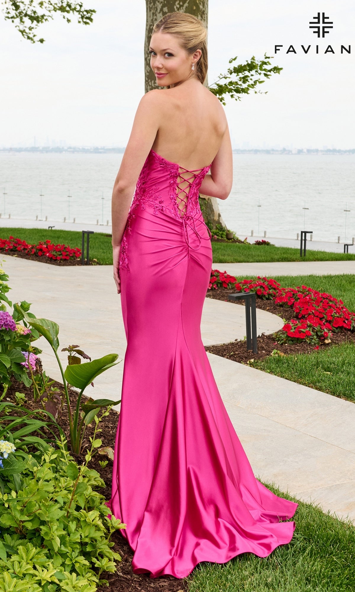 A woman wears a sleeveless, hot pink mermaid-style Faviana 11647 prom dress with floral beaded appliques and a lace-up back, standing on a garden path near the water.
