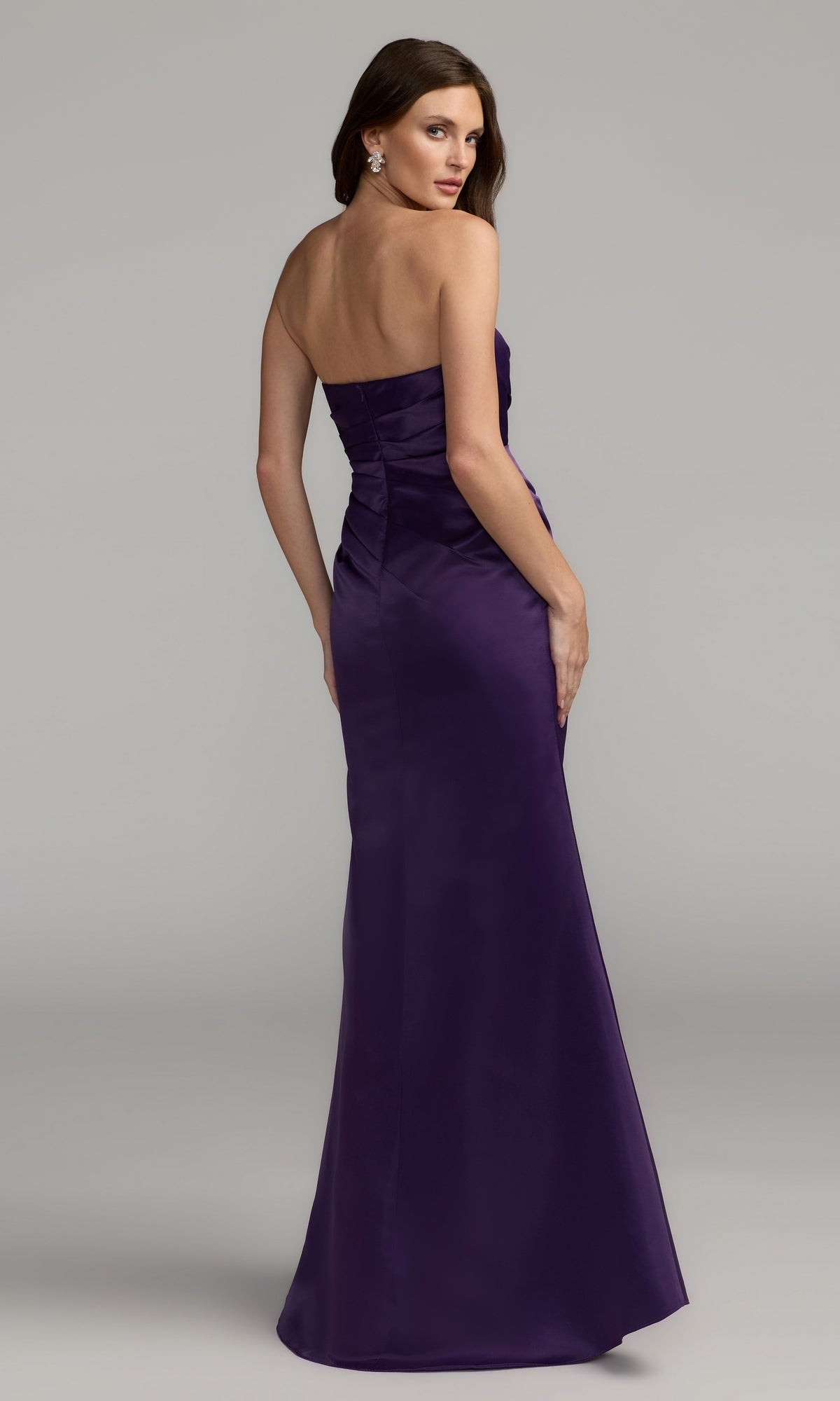 A woman models the Strapless Long Prom Dress: Mac Duggal 11653, standing with her back slightly turned to the camera in front of a plain gray background. The floor-length, fitted purple gown elegantly accentuates her silhouette, offering a sophisticated and timeless appearance.