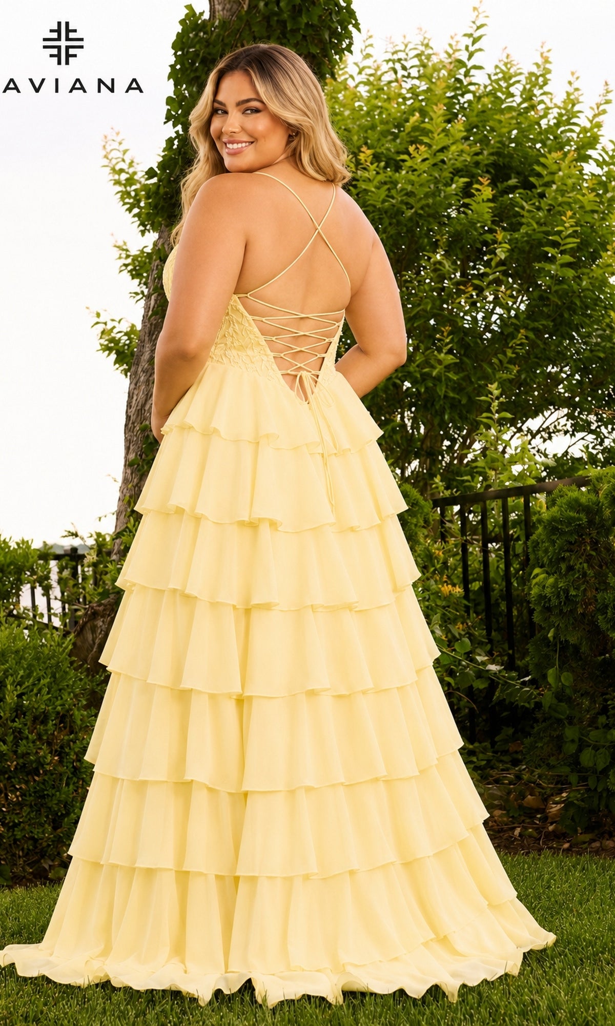 A woman stands outdoors wearing the Long Plus-Size Prom Dress: Faviana 11665E, a floor-length, light yellow chiffon ruffle dress with a tiered skirt and a lace-up corset back.