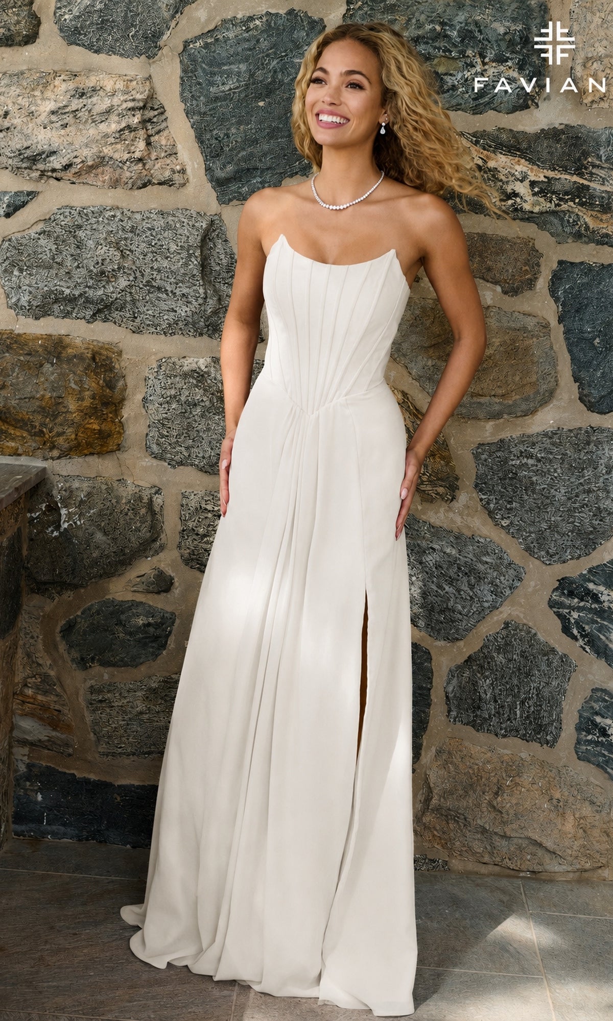 A woman wearing an ivory, strapless Faviana 11672 corset prom dress with a floor-length, side-slit skirt, standing in front of a stone wall.