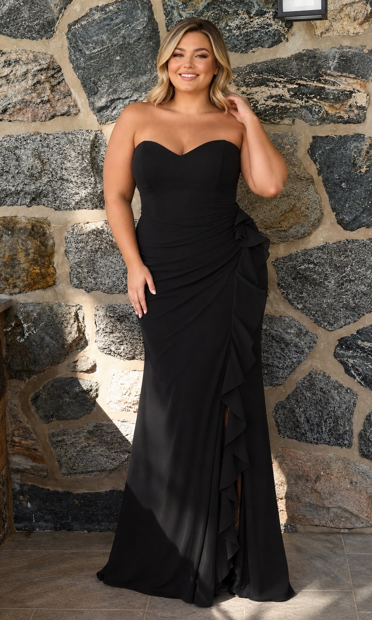 A woman wears a black, strapless, floor-length Long Plus-Size Prom Dress: Faviana 11673E with a sweetheart neckline and a ruffled side slit.