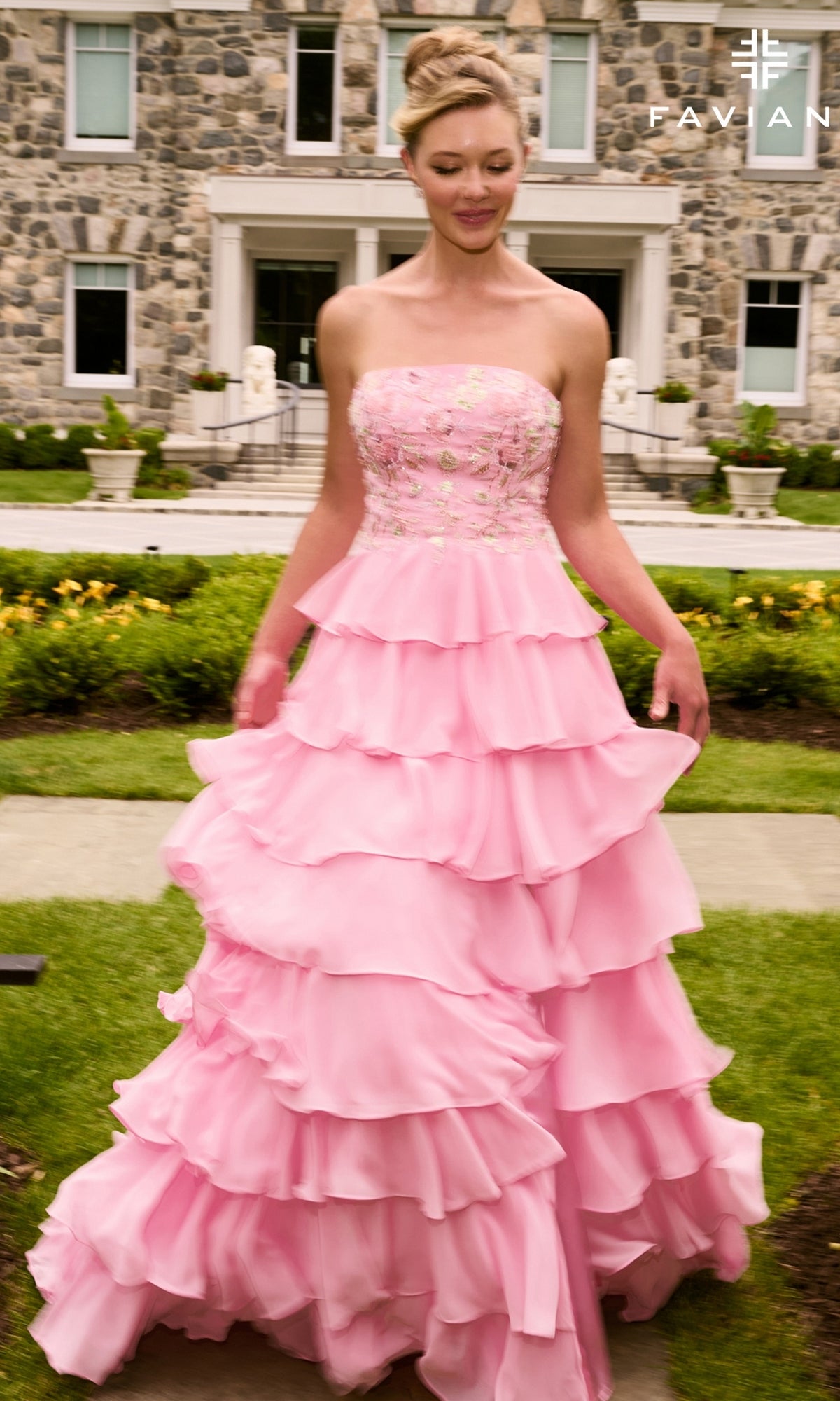 A person wears a Faviana 11675 long prom dress, a strapless pink gown with floral applique, an embroidered bodice, and a tiered skirt, standing on a lawn in front of a stone building.