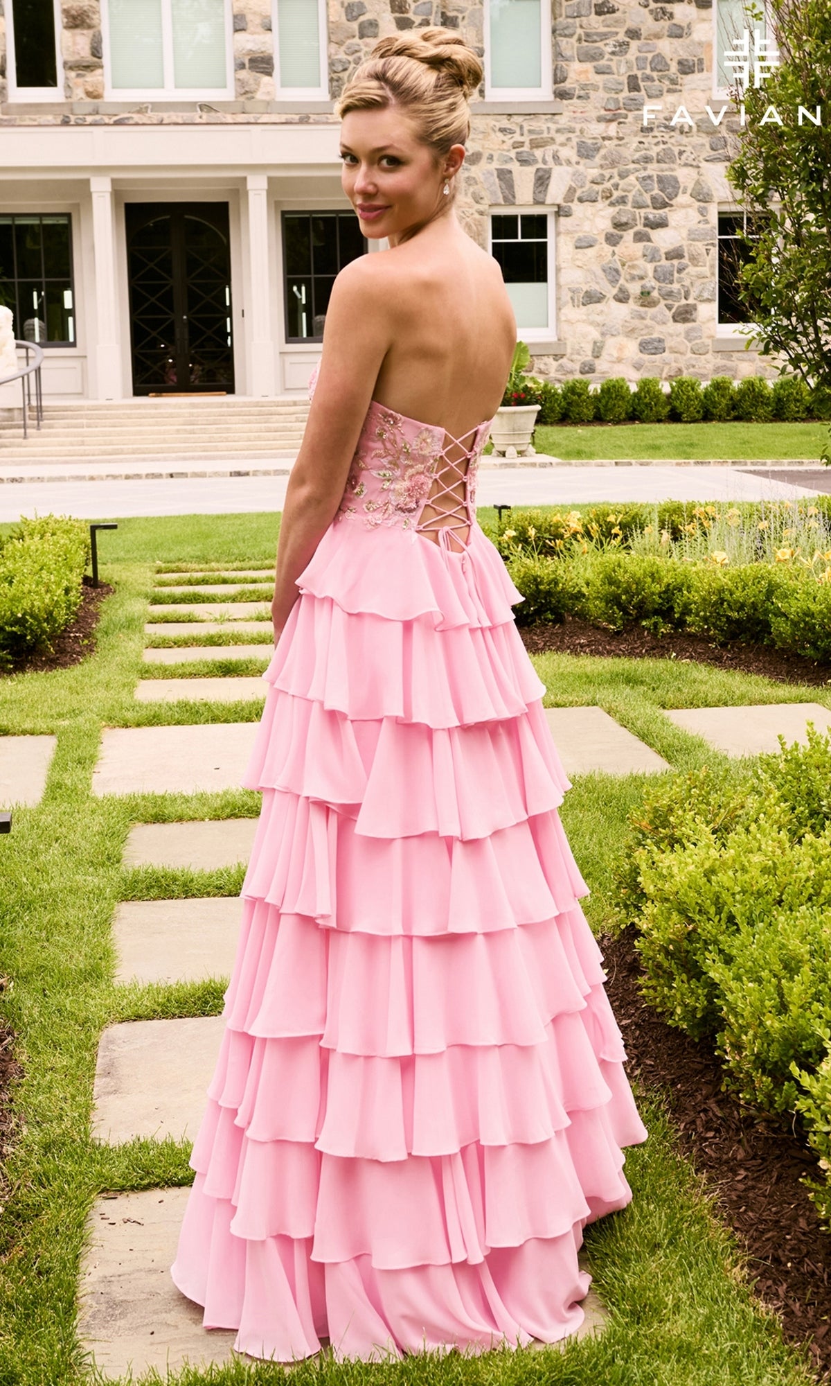A woman stands on a stone garden path wearing a Faviana 11675, a strapless, floor-length pink gown with a lace-up back and a tiered ruffle skirt.