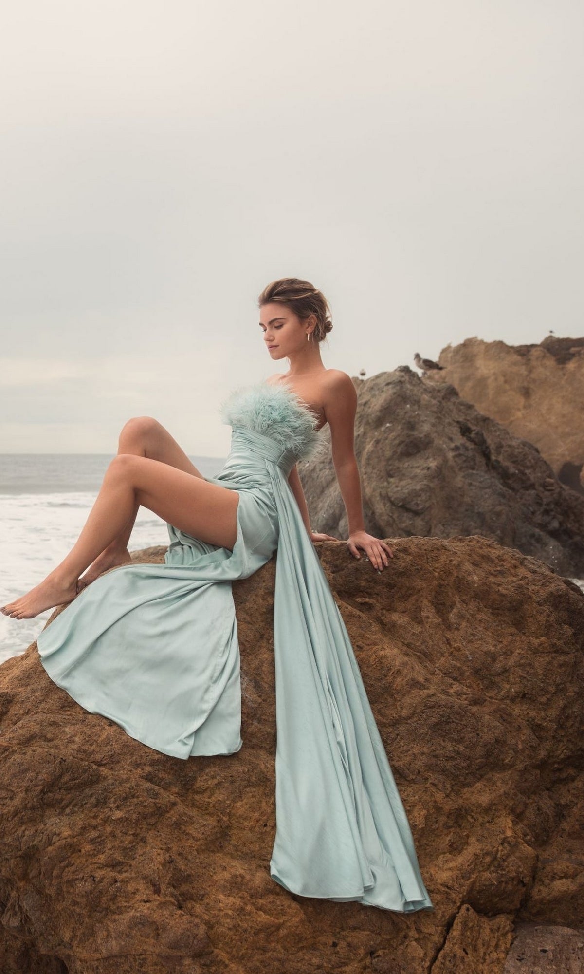 A woman wearing the Long Formal Dress 11690 by Mac Duggal—a light blue satin strapless gown with a high slit—sits on a large rock by the ocean, cliffs and a cloudy sky in the background.