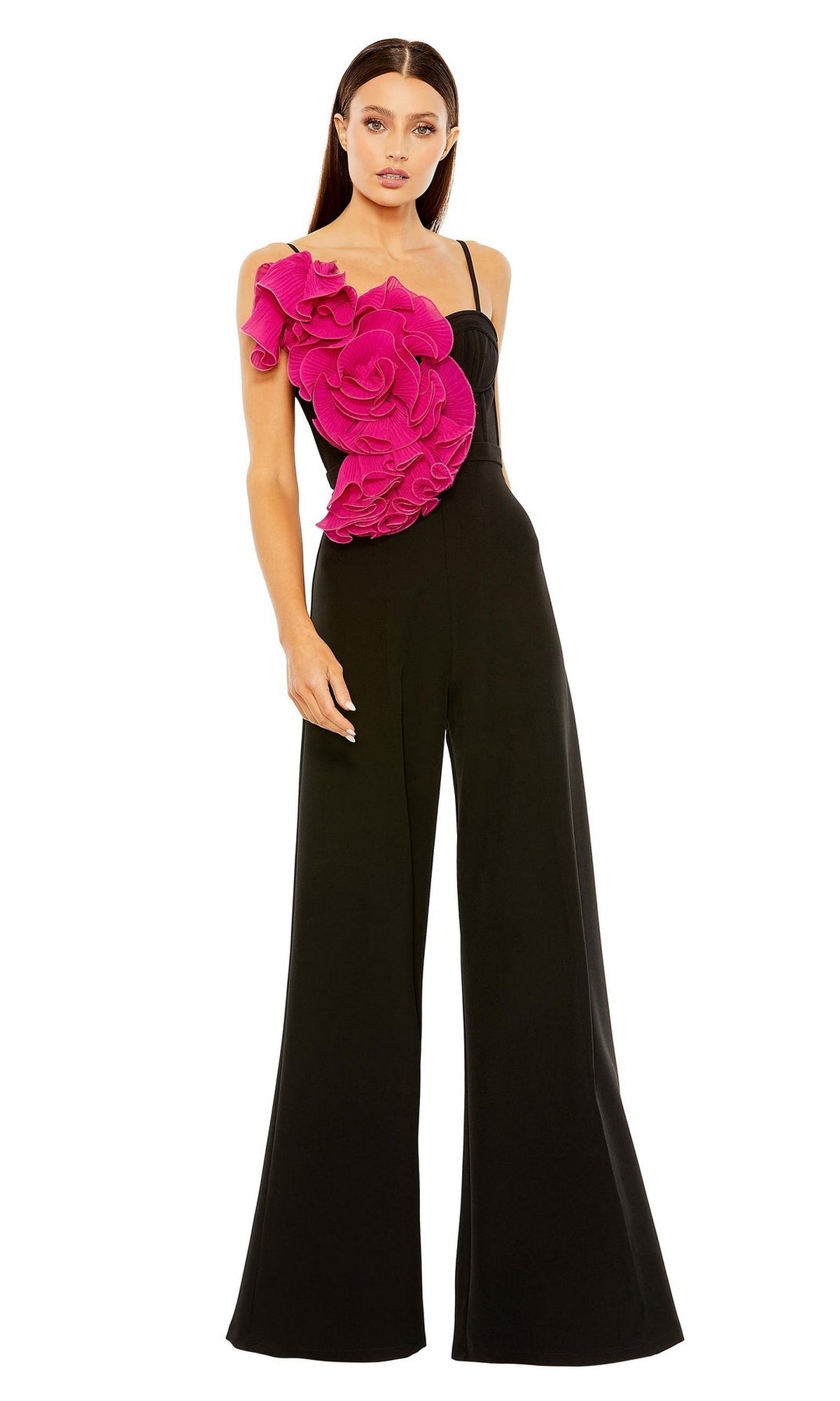 A woman stands against a white background wearing the Long Formal Jumpsuit 11767 by Mac Duggal, featuring black wide-leg pants and a large bright pink floral applique on one shoulder.