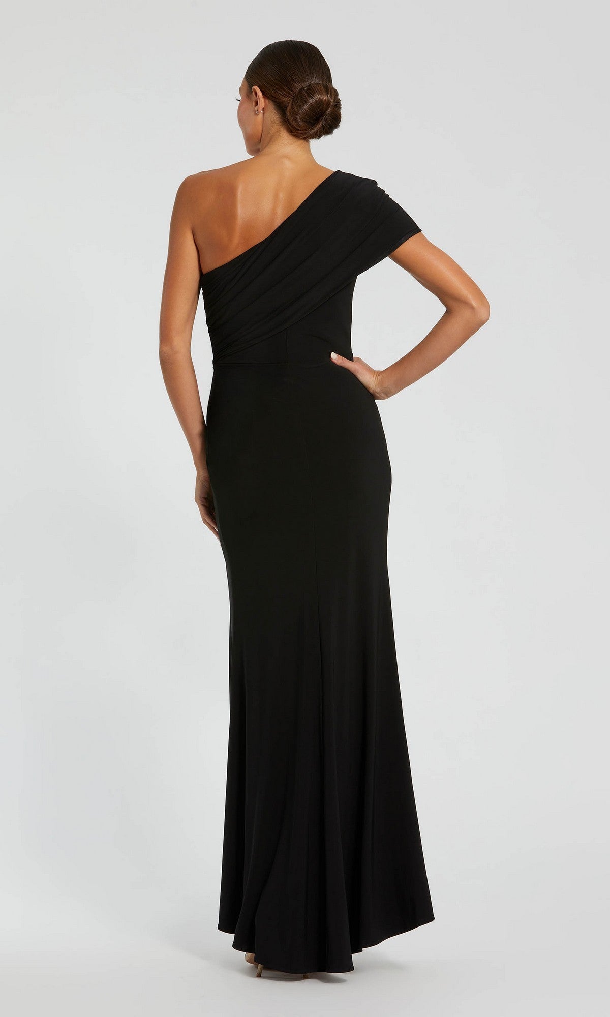 A woman stands with her back to the camera, wearing the Mac Duggal 11769 long formal dress, featuring a fitted one-shoulder jersey design, against a plain light background.