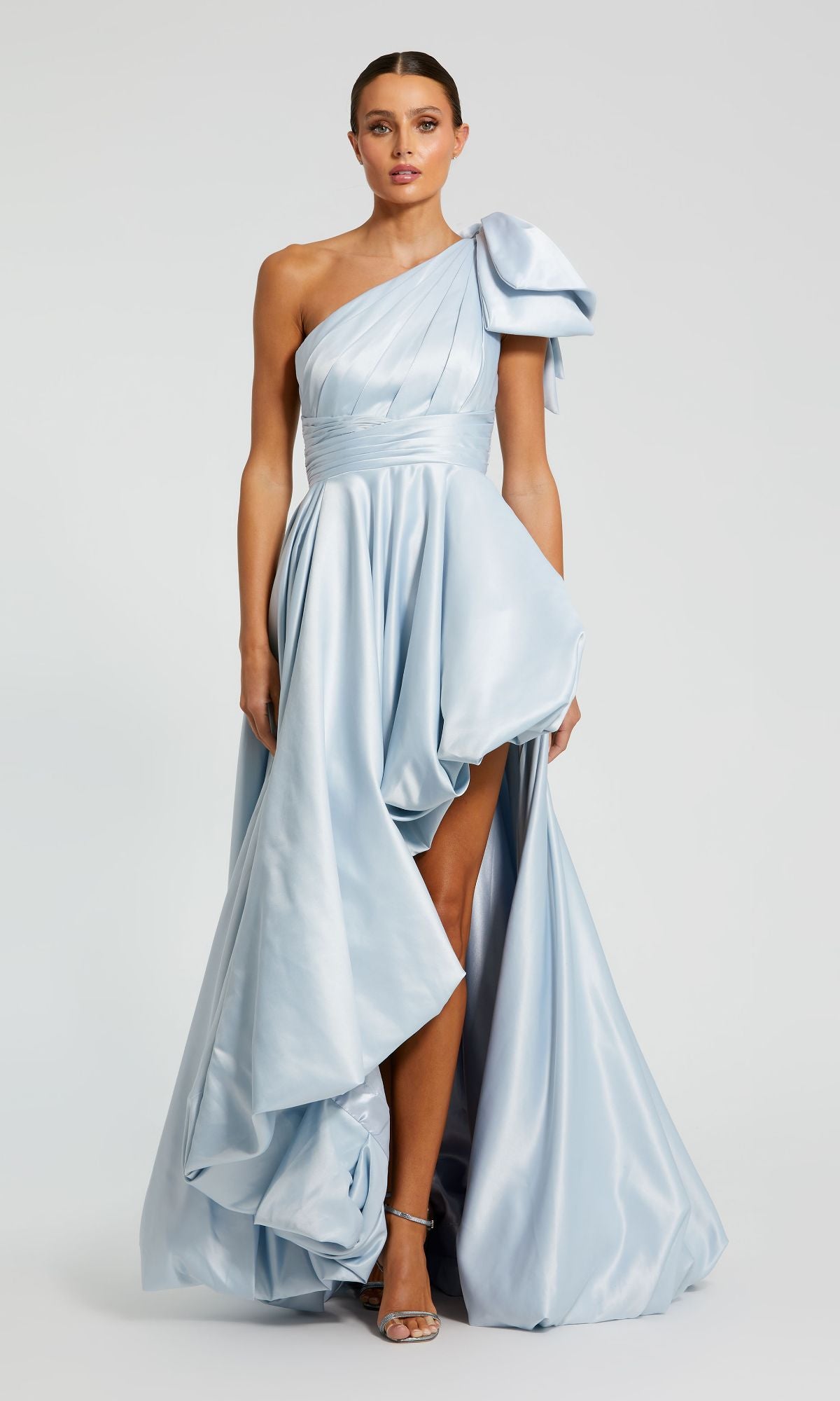 A woman models the Bow Long Bubble Prom Dress: Mac Duggal 11800—light blue, one-shoulder style with draped details, asymmetrical hem, and high slit—standing against a plain background.